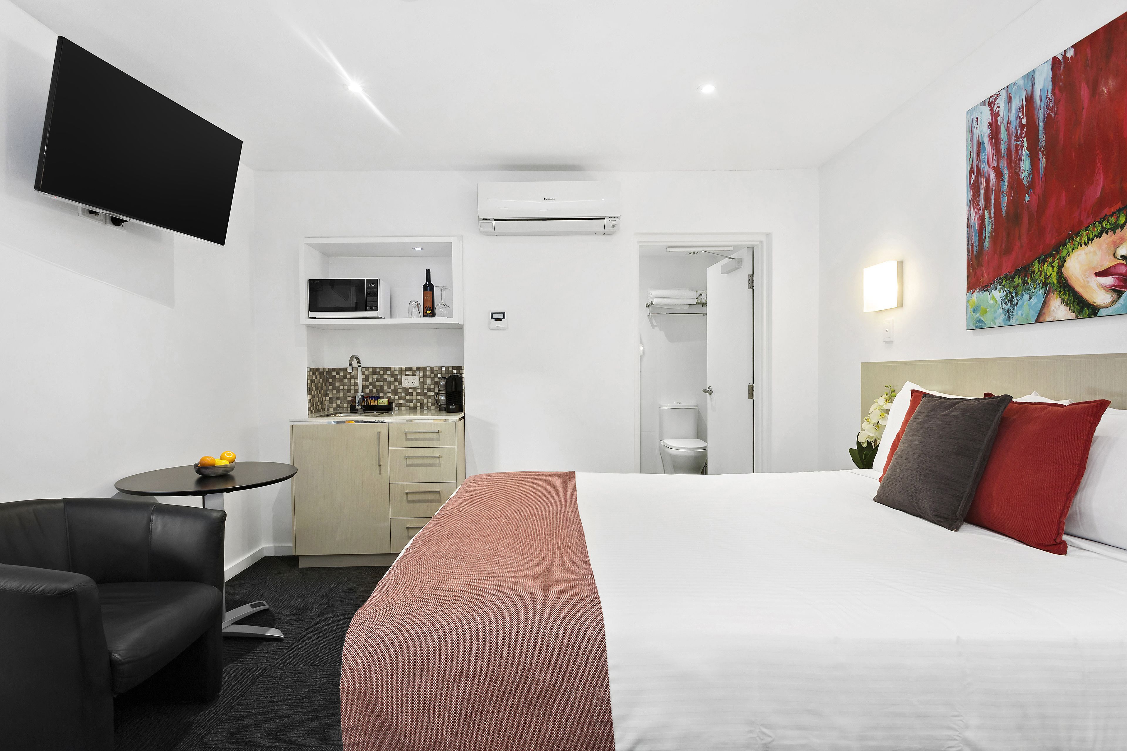 A hotel room with a bed, a TV, a small kitchenette, a chair, and a painting on the wall.