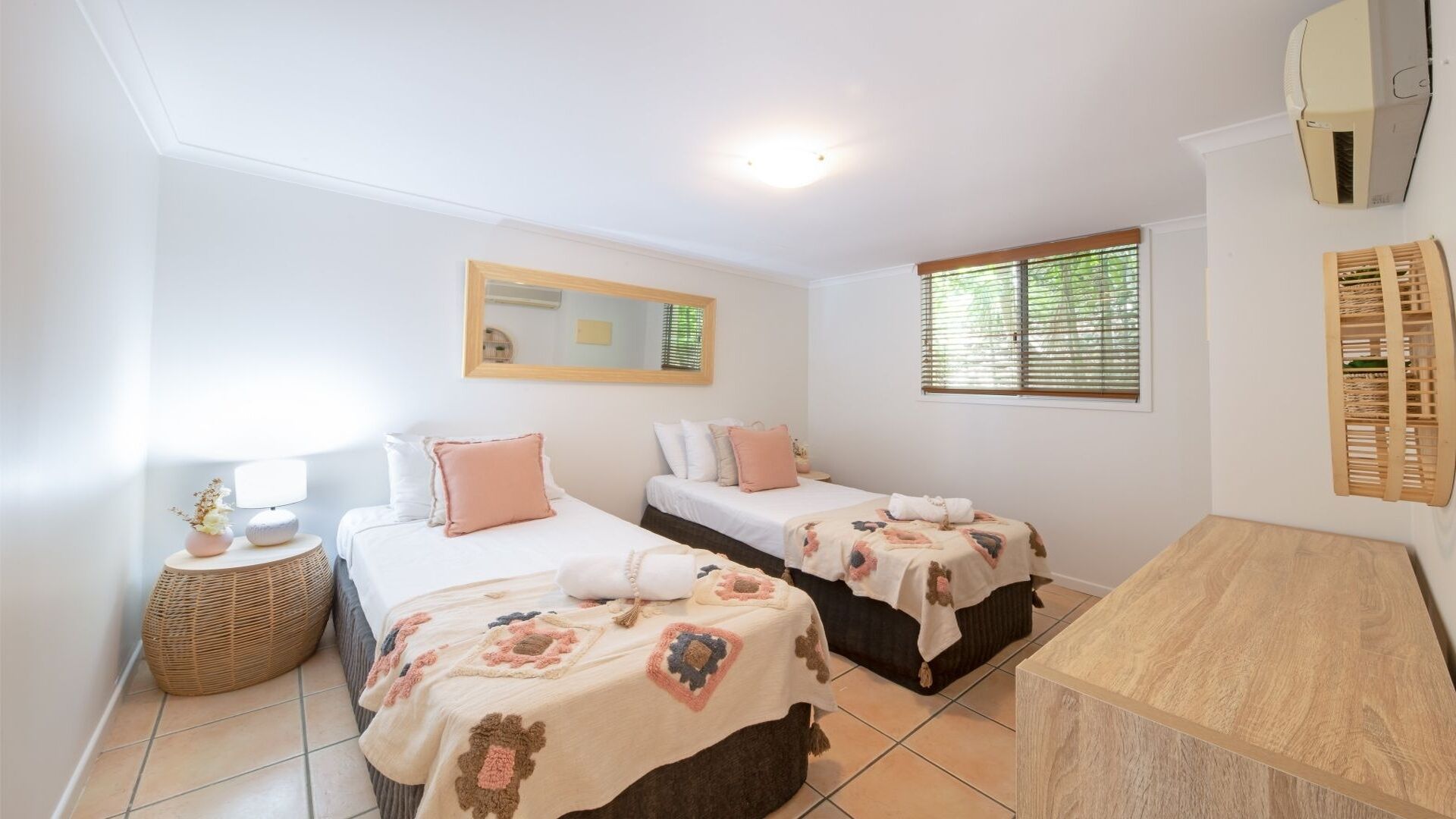 Un dormitorio con dos camas, una mesita de noche con lámpara, una pequeña mesa redonda con una cesta y un aparador de madera.