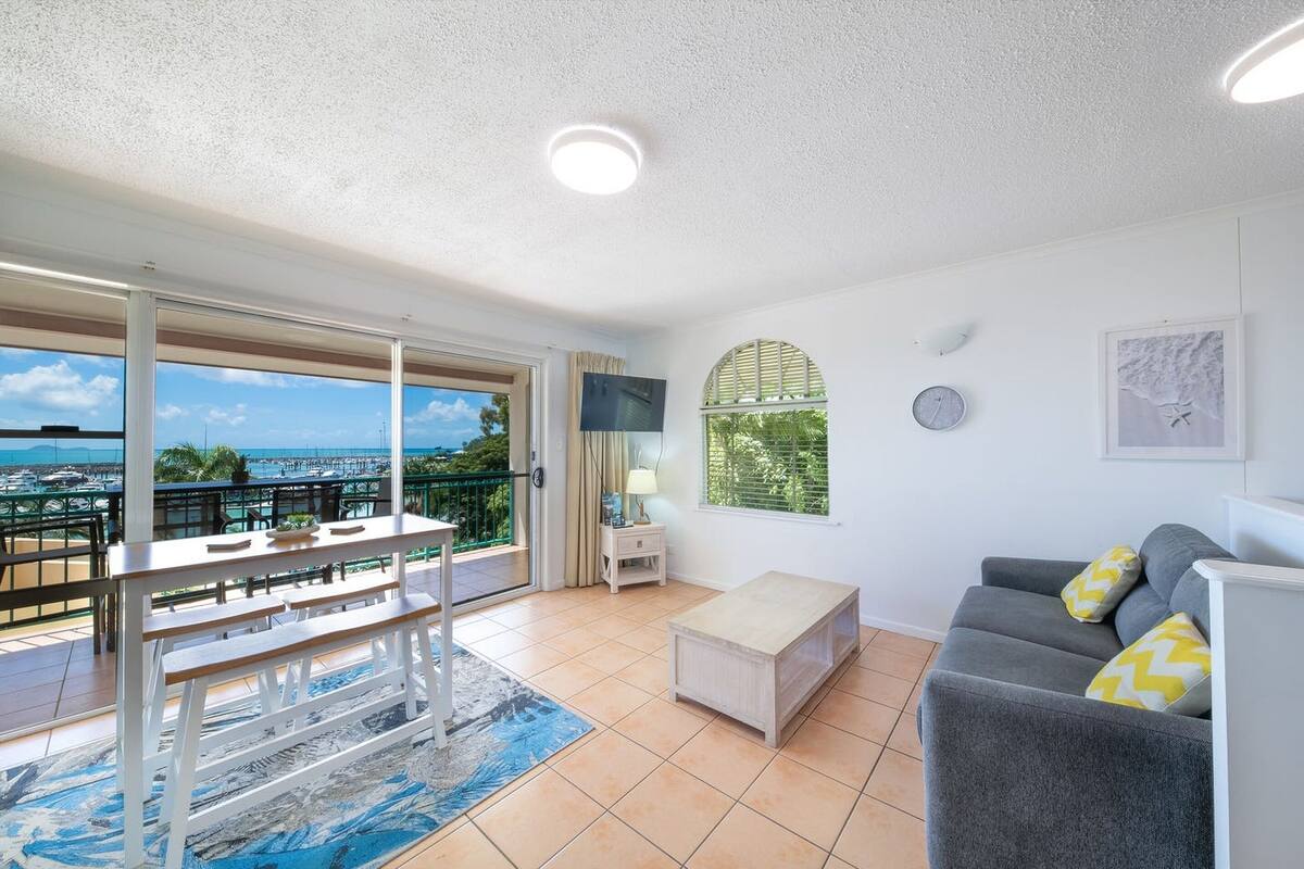 A modern living room with a balcony overlooking a marina, a grey sofa, a wooden bench, and a small table with a lamp.