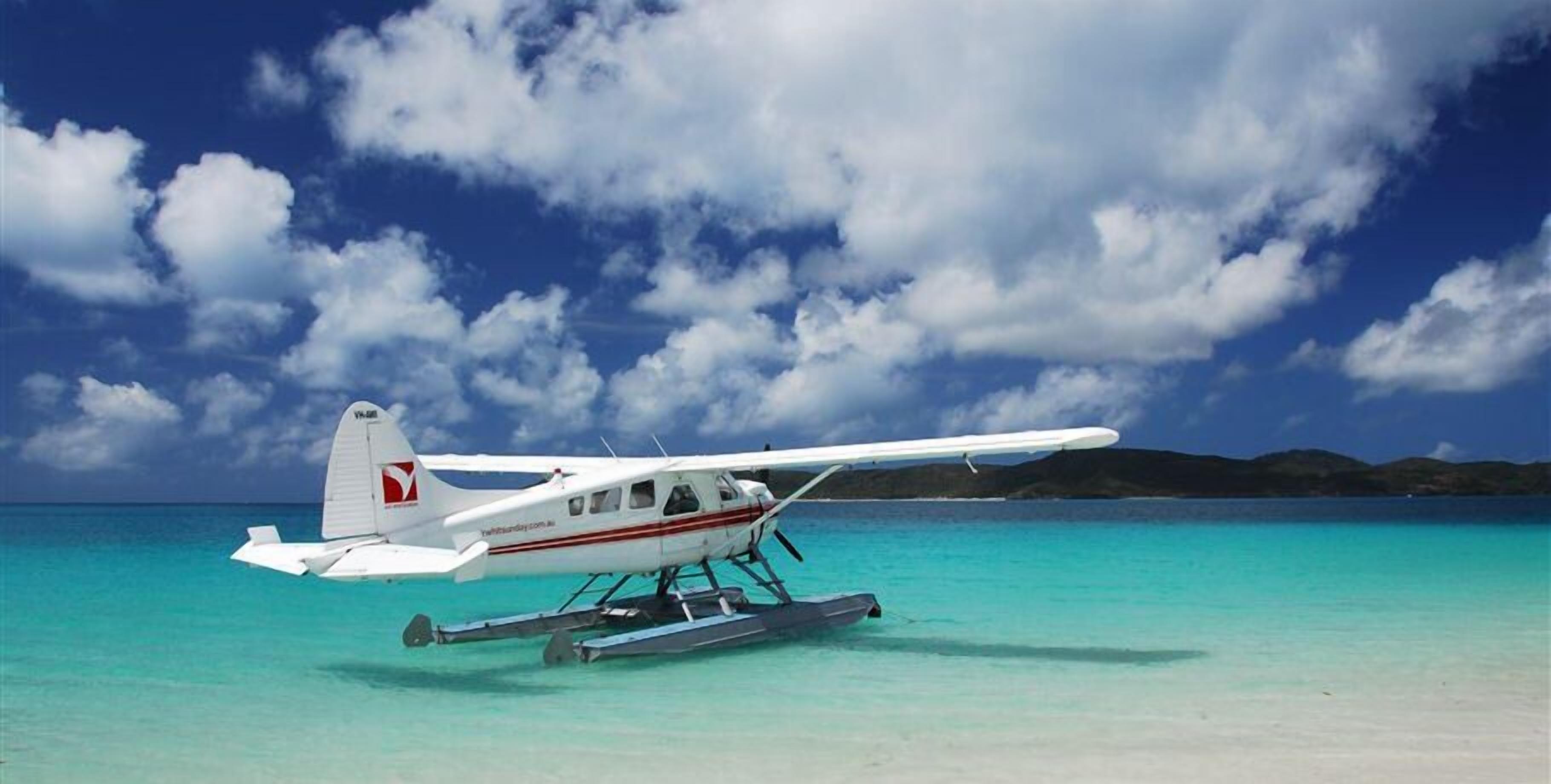 A seaplane parked on a beach with clear turquoise water and a mountainous backdrop.