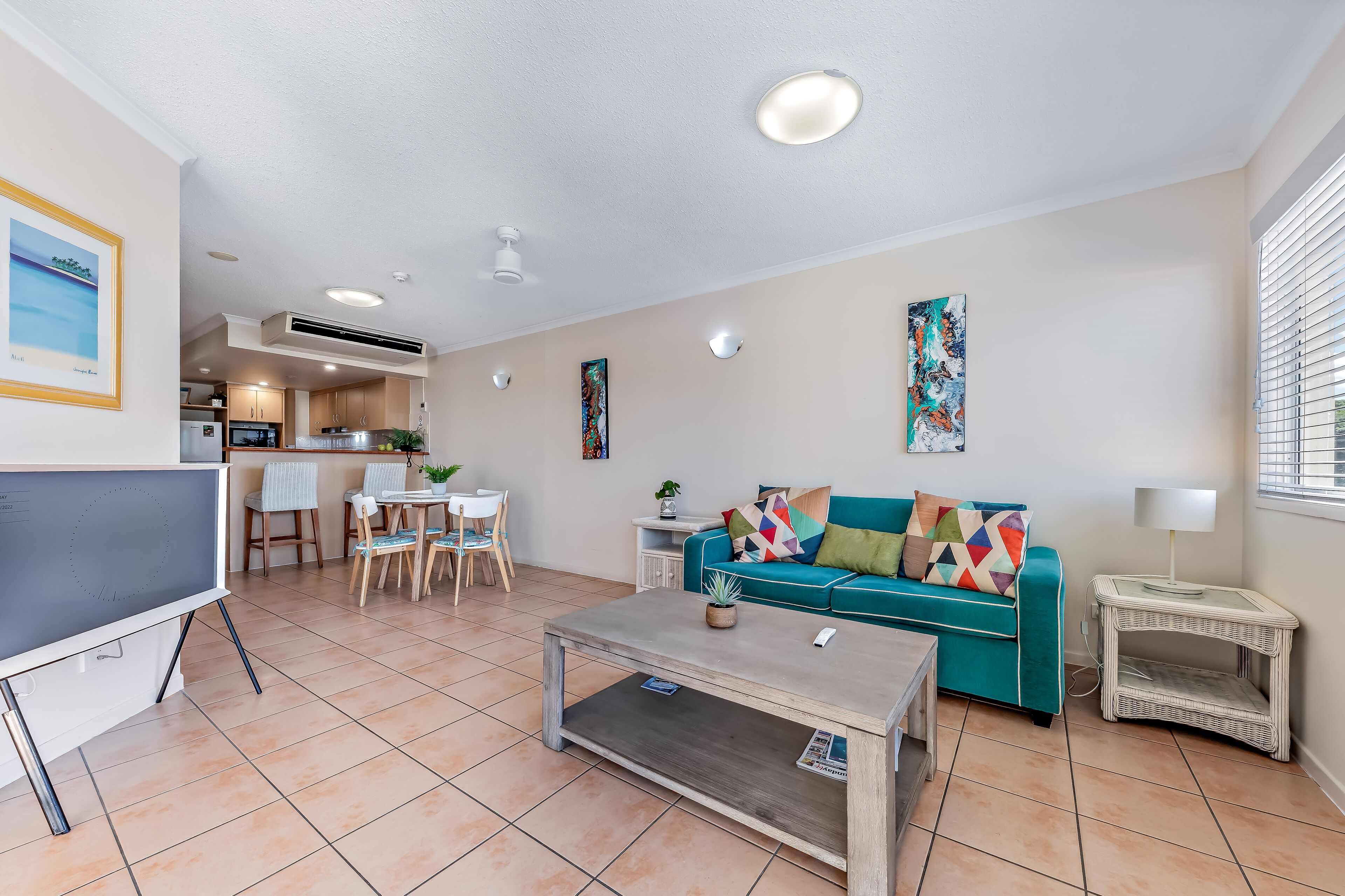 Una sala moderna con un sofá color turquesa, una mesa de centro y un televisor. Se observa un comedor con sillas y una mesa, y una cocina al fondo.