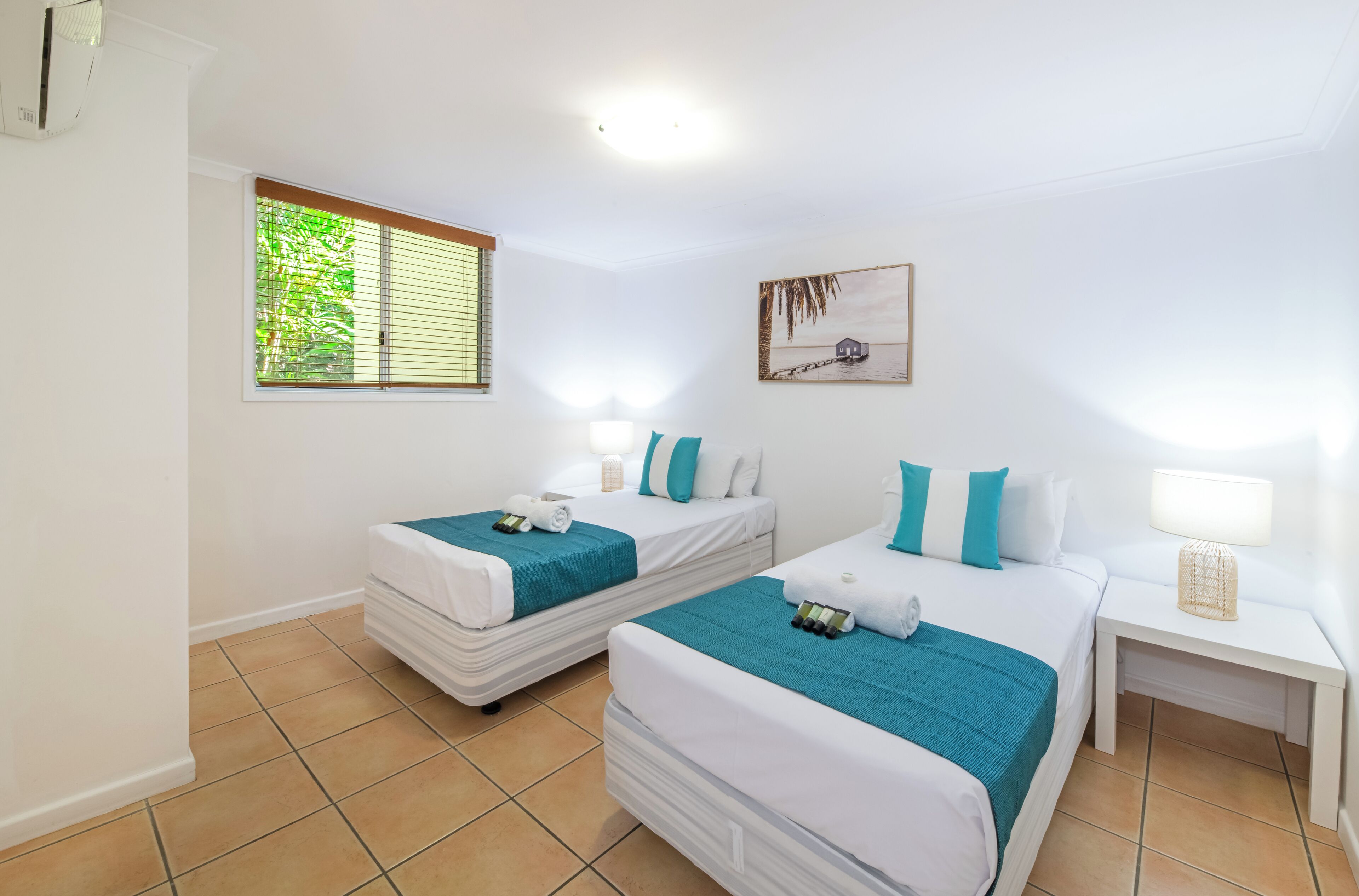 A hotel room with two beds, a window with blinds, a painting on the wall, and a bedside table with a lamp.