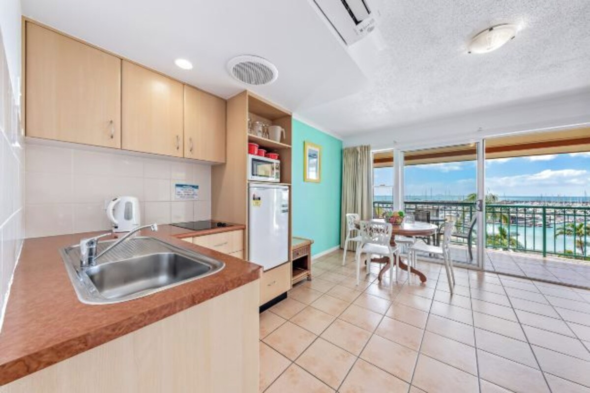 A modern kitchen with a sink, cabinets, and a refrigerator. There is a dining area with a table and chairs, and a balcony with a view of the sea.