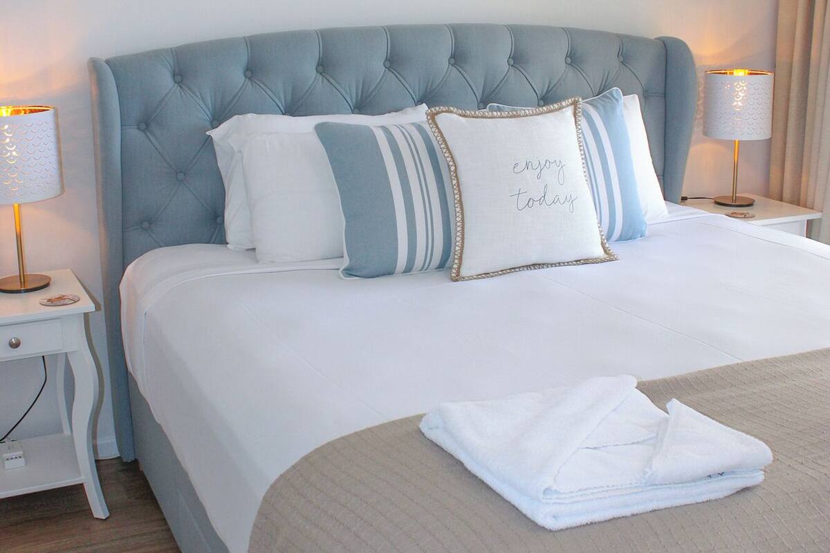 A neatly made bed with a tufted headboard, white bedding, and a decorative pillow with text. There are two bedside tables with lamps, and wall art above the bed.