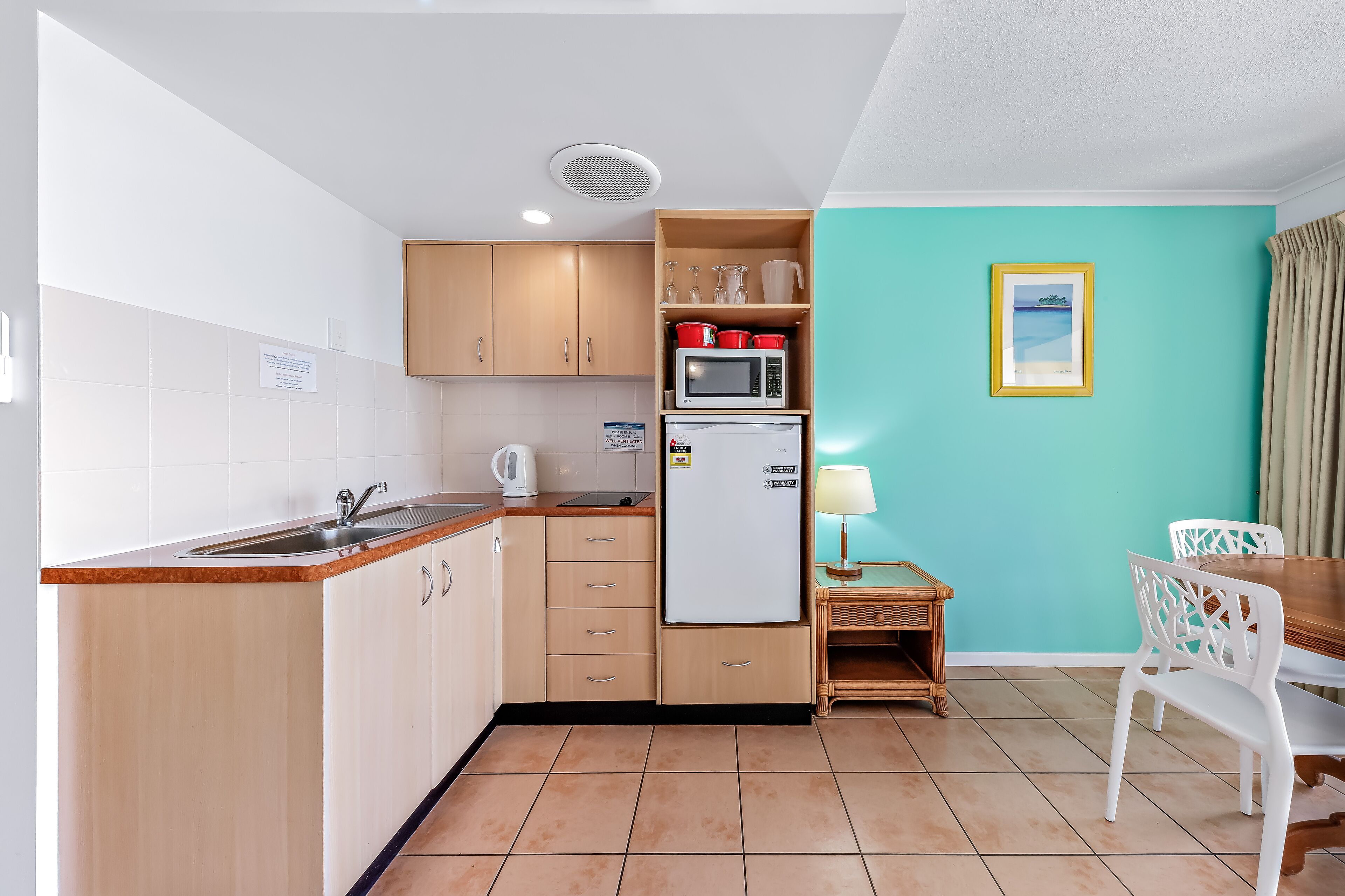 A compact kitchen with a sink, microwave, and refrigerator, a dining table with chairs, and a teal wall with a framed picture.