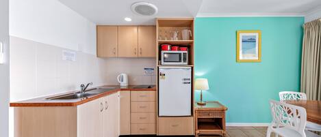 Cocina compacta con lavamanos, microondas y refrigerador, una mesa de comedor con sillas, y una pared verde azulado con un cuadro enmarcado.