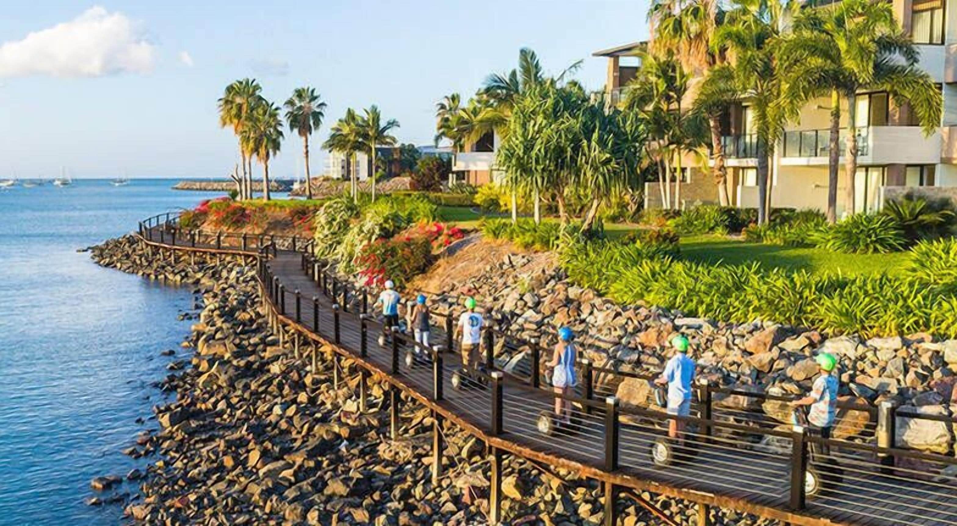 Une promenade en bord de mer avec une terrasse en bois, des palmiers et une côte rocheuse.