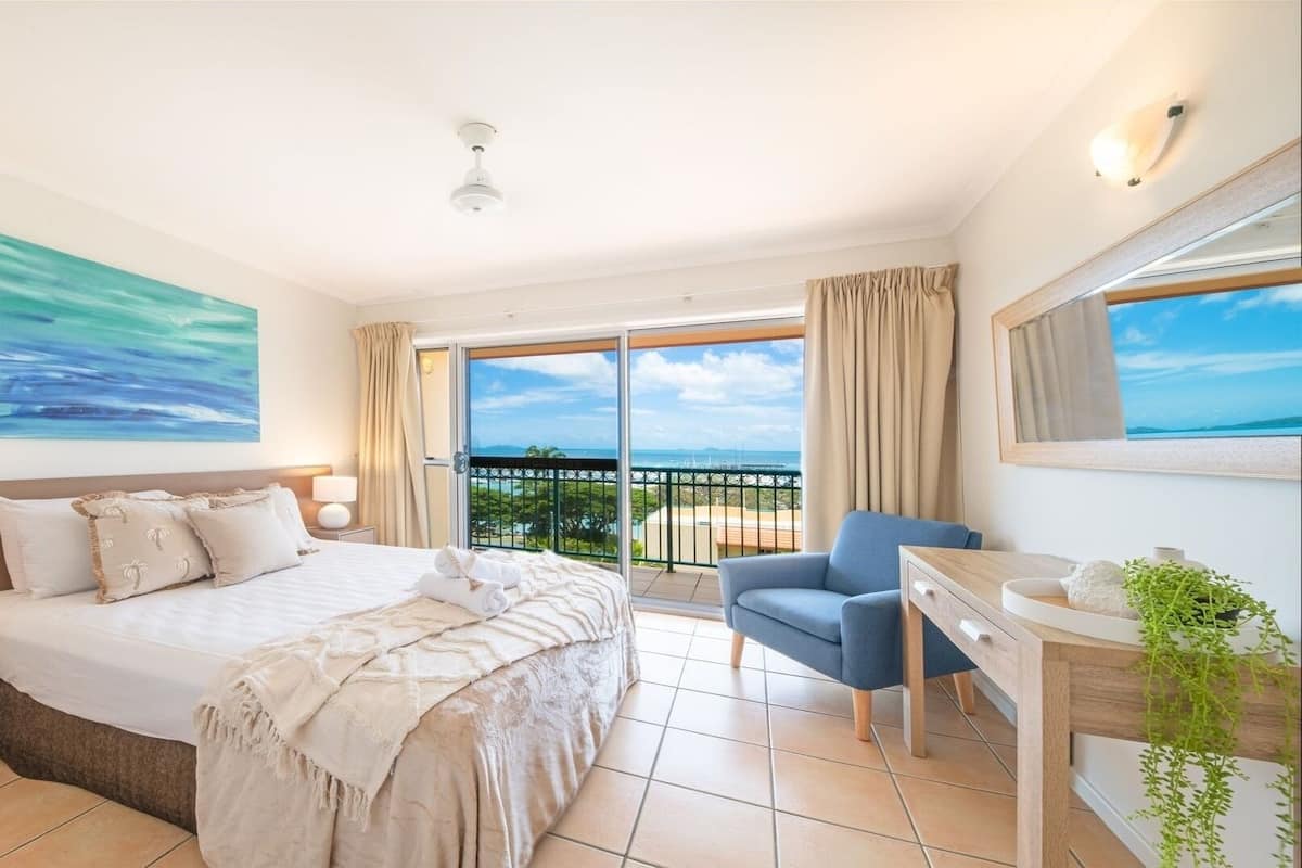 A hotel room with a bed, a desk, a chair, a balcony with a view of the sea, and a painting on the wall.