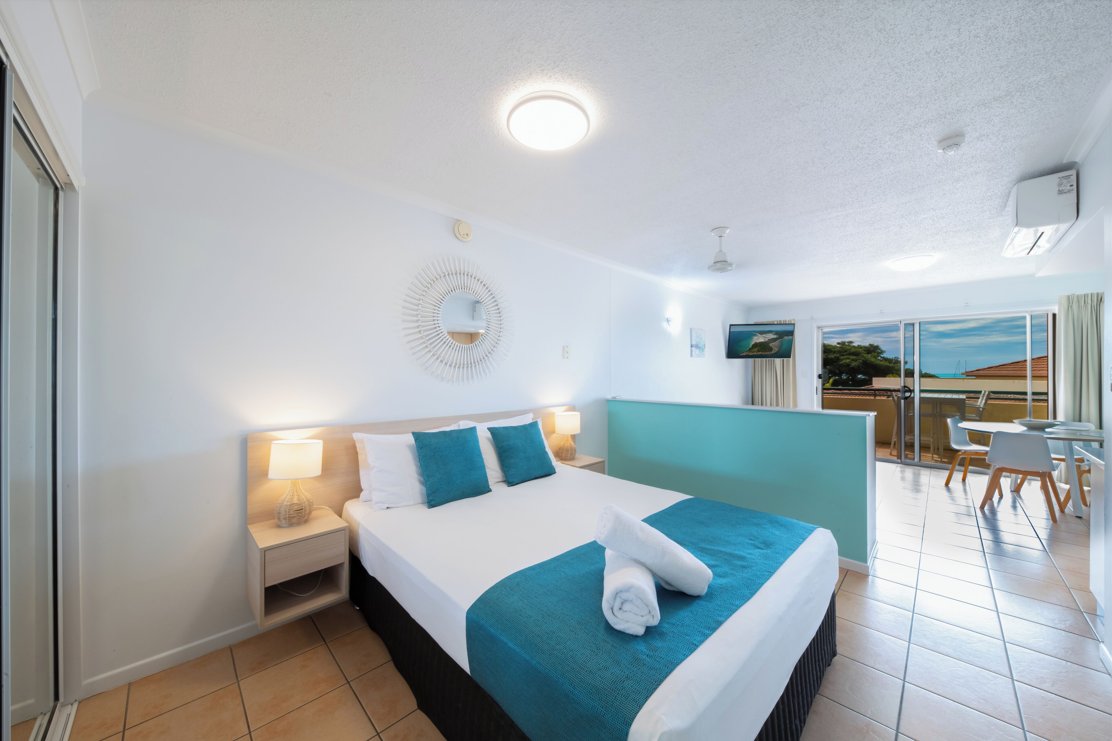 A hotel room with a bed, a nightstand, a wall-mounted lamp, a ceiling light, a TV, and a balcony with a table and chairs.