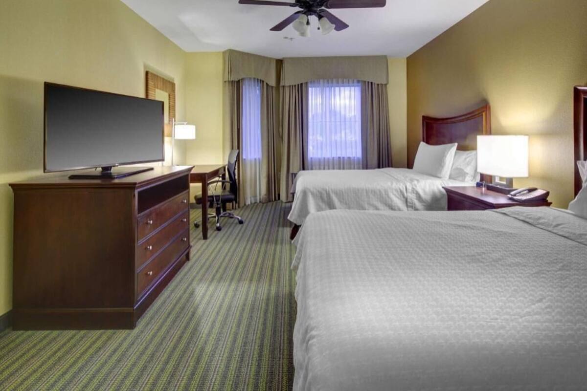 Habitación de hotel con dos camas, televisión, escritorio y ventilador de techo.