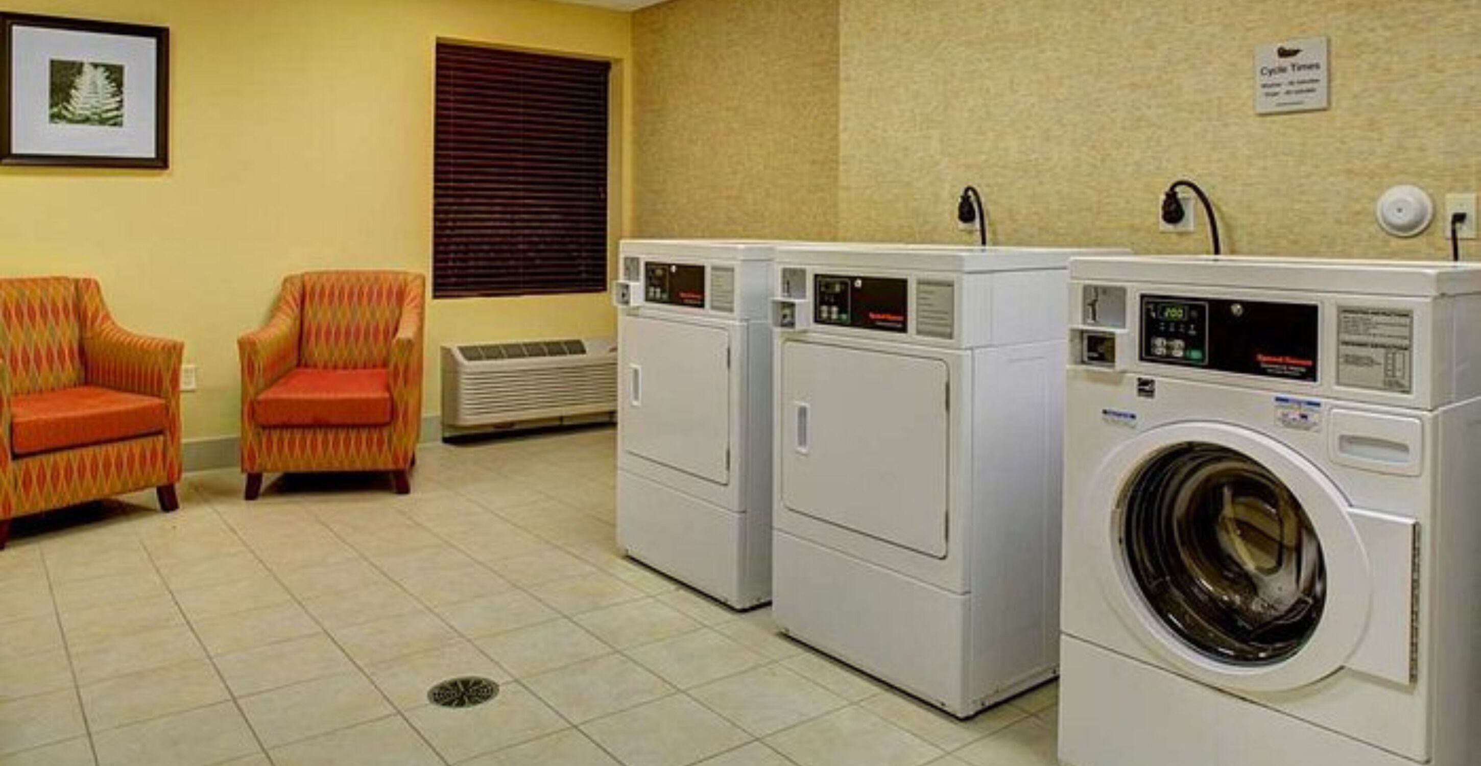 A laundry room with two dryers, a washing machine, and two patterned chairs.