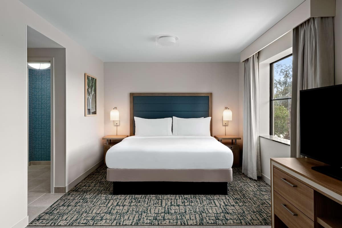 Una habitación de hotel con una cama grande, dos lámparas de noche, un televisor de pantalla plana, un aparador de madera y una ventana con cortinas.