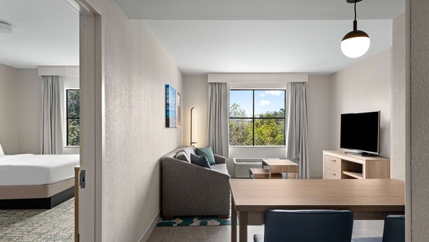A hotel room with a bed, a sofa, a dining table with chairs, a TV, and a window with curtains. - Homewood Suites West Palm Beach (West Palm Beach)