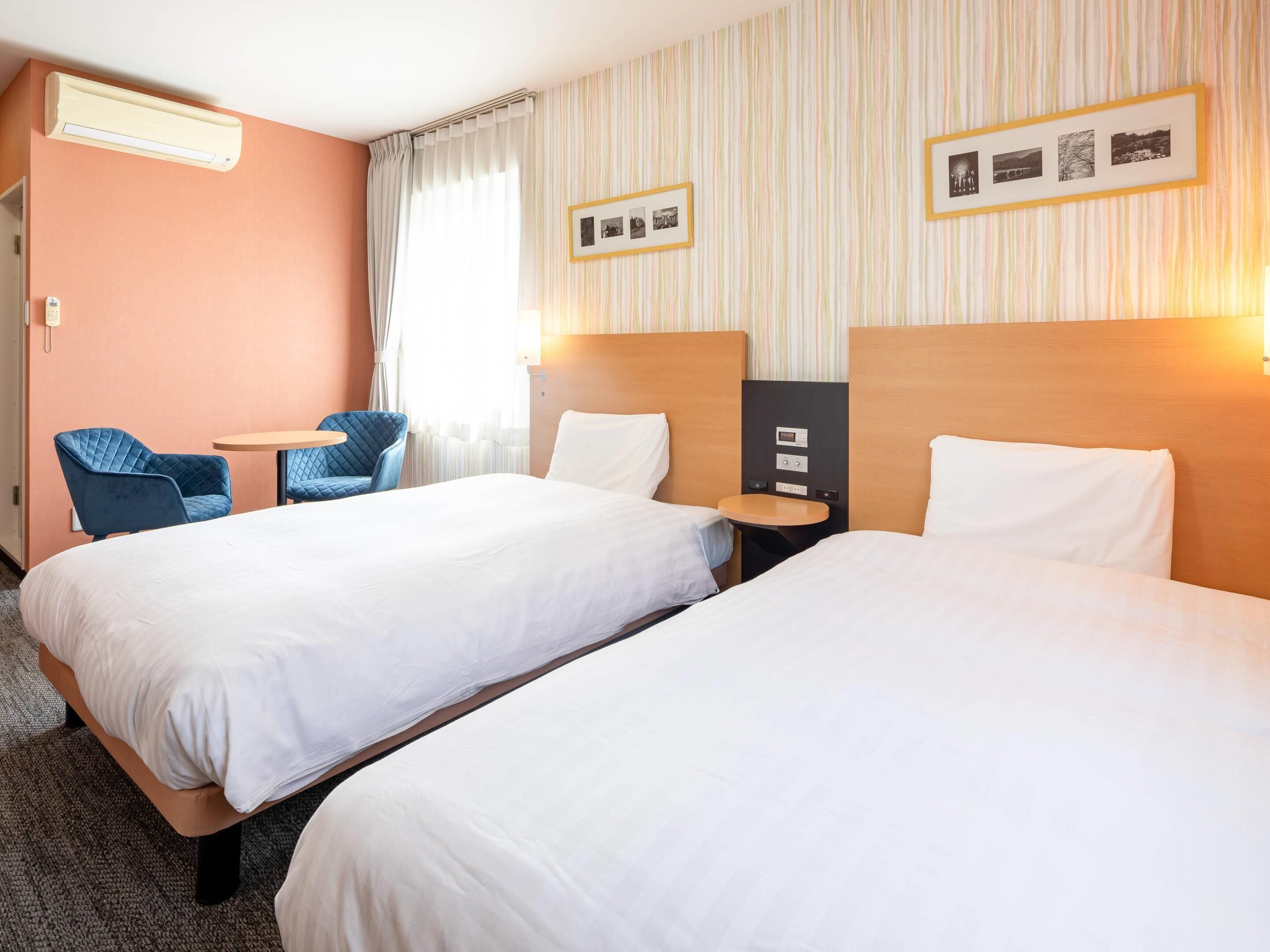A hotel room with two beds, a small table, a chair, and a wall-mounted air conditioner.