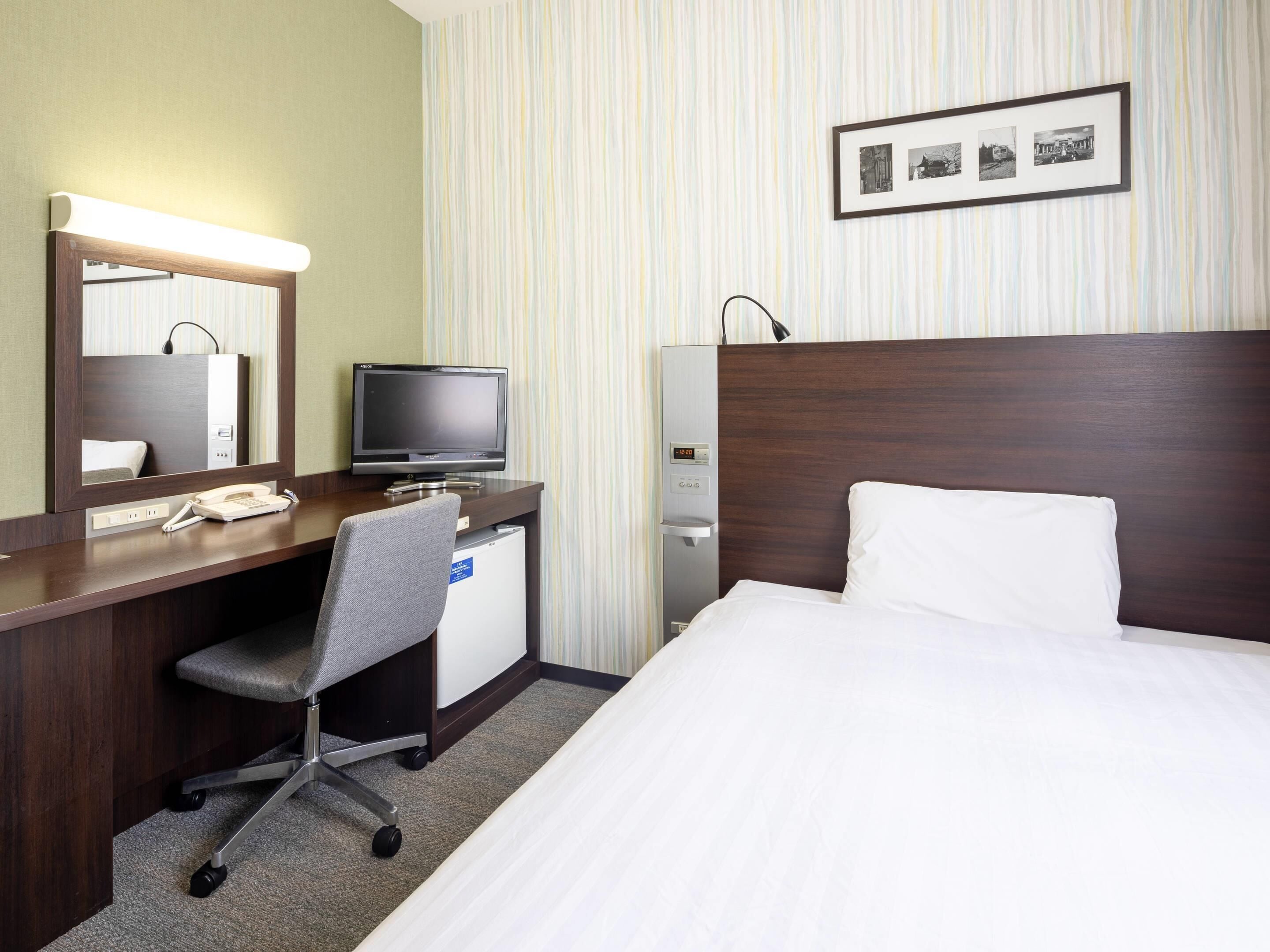 Hotelkamer met een bed, een bureau met een computer, een telefoon, een spiegel, een wandklok en een ingelijste foto.