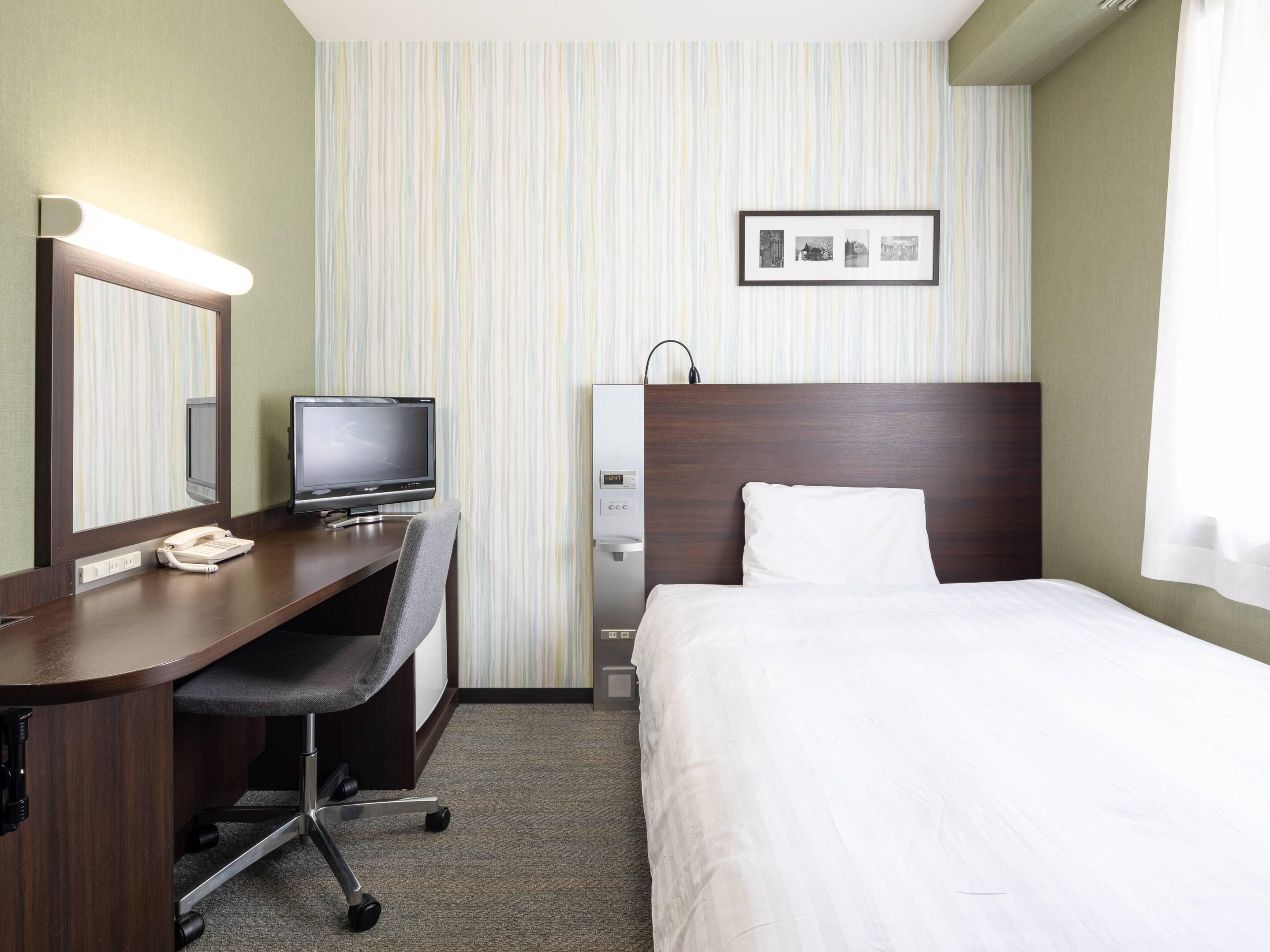 A hotel room with a bed, a desk with a computer and phone, a mirror, a wall-mounted TV, and a picture frame.