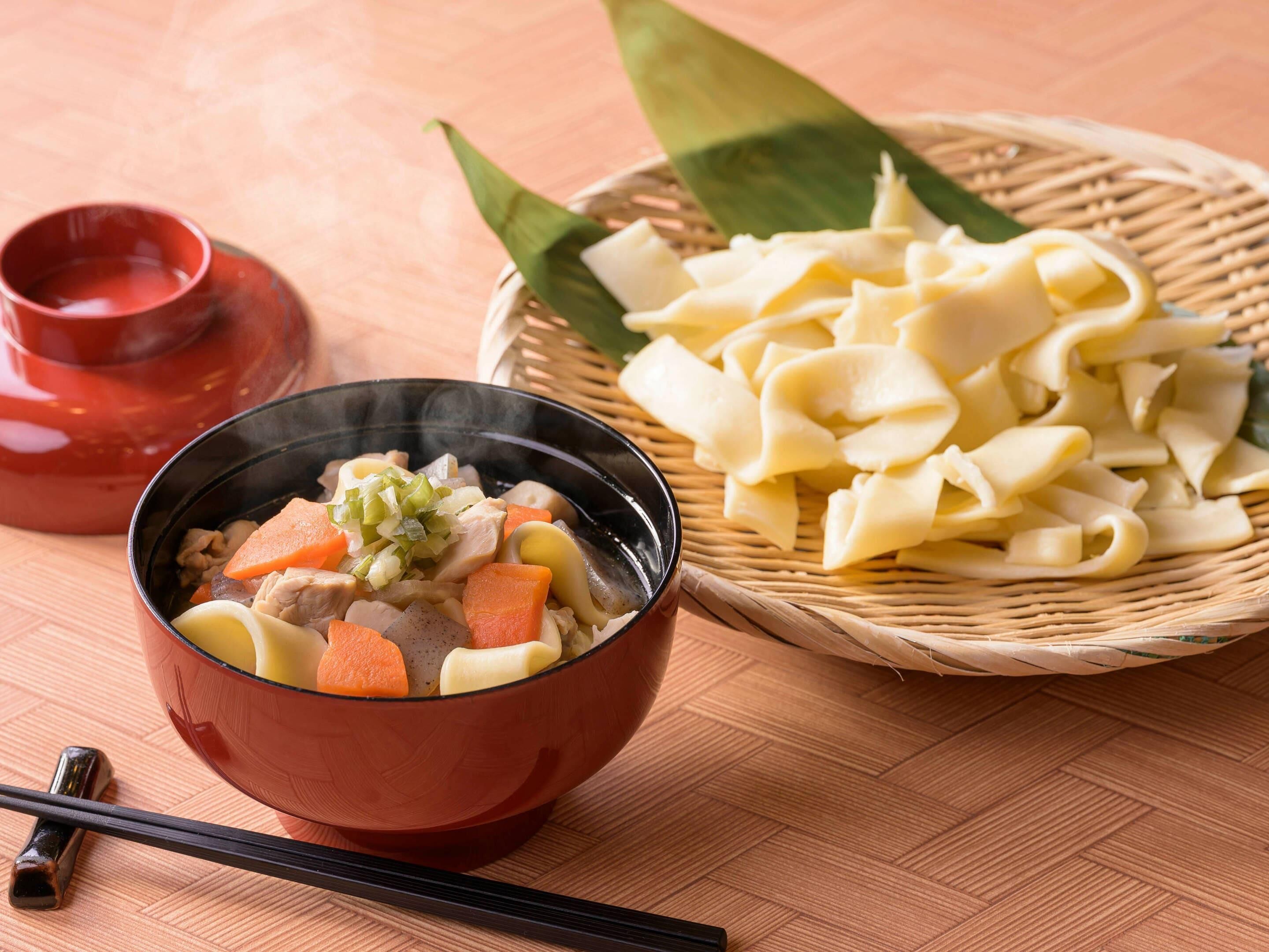 A bowl of soup with vegetables and meat, a basket of noodles, and chopsticks on a wooden table.