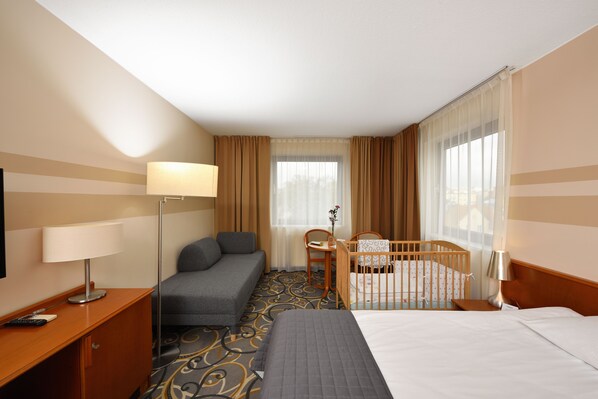 A hotel room with a bed, a desk with a lamp, a sofa, a crib, and a window with curtains.