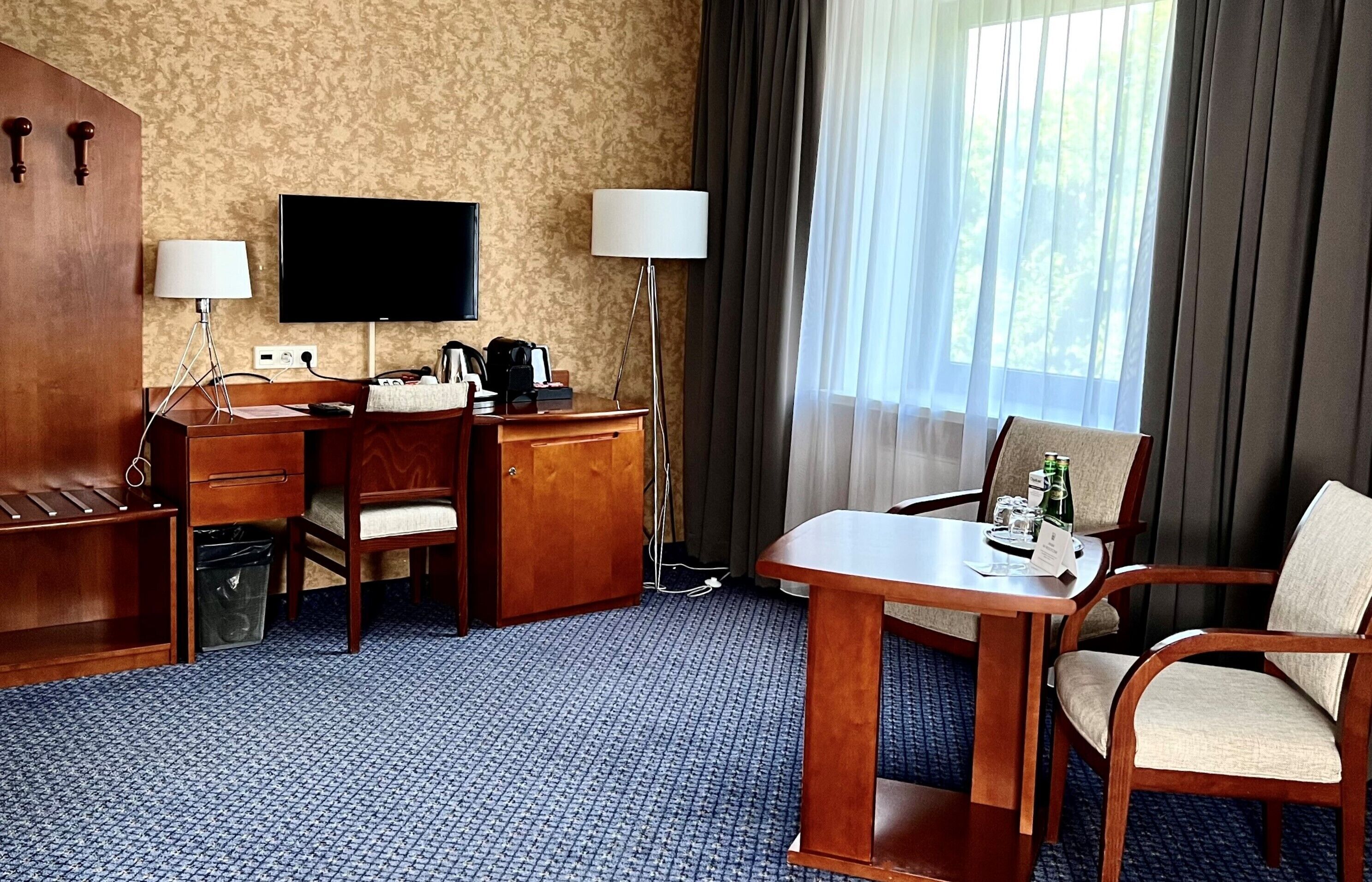 Hotelkamer met een houten bureau, twee stoelen, een televisie, een lamp en uitzicht op het landschap door een raam met gordijnen.