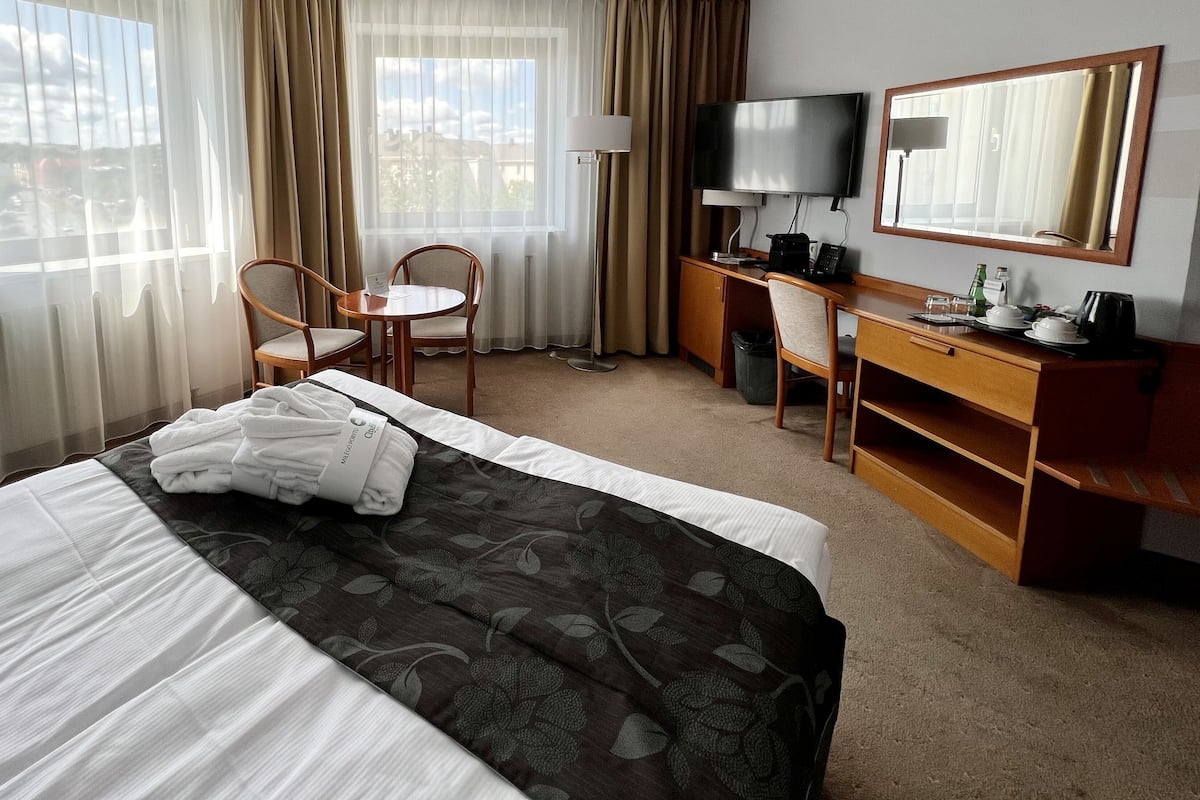Habitación de hotel con cama, escritorio, silla, televisor y una ventana grande con cortinas.