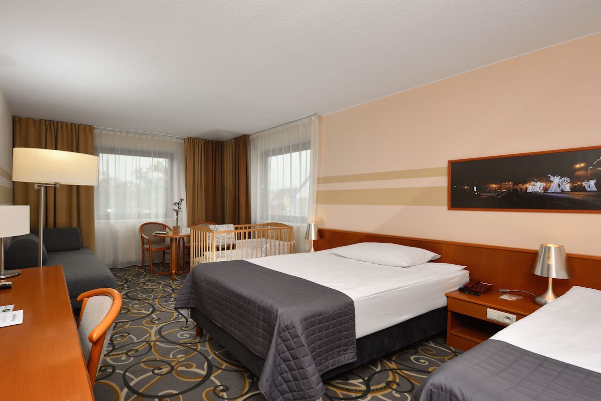 Habitación de hotel con dos camas, un escritorio con lámpara, una silla, una cuna y un cuadro en la pared.