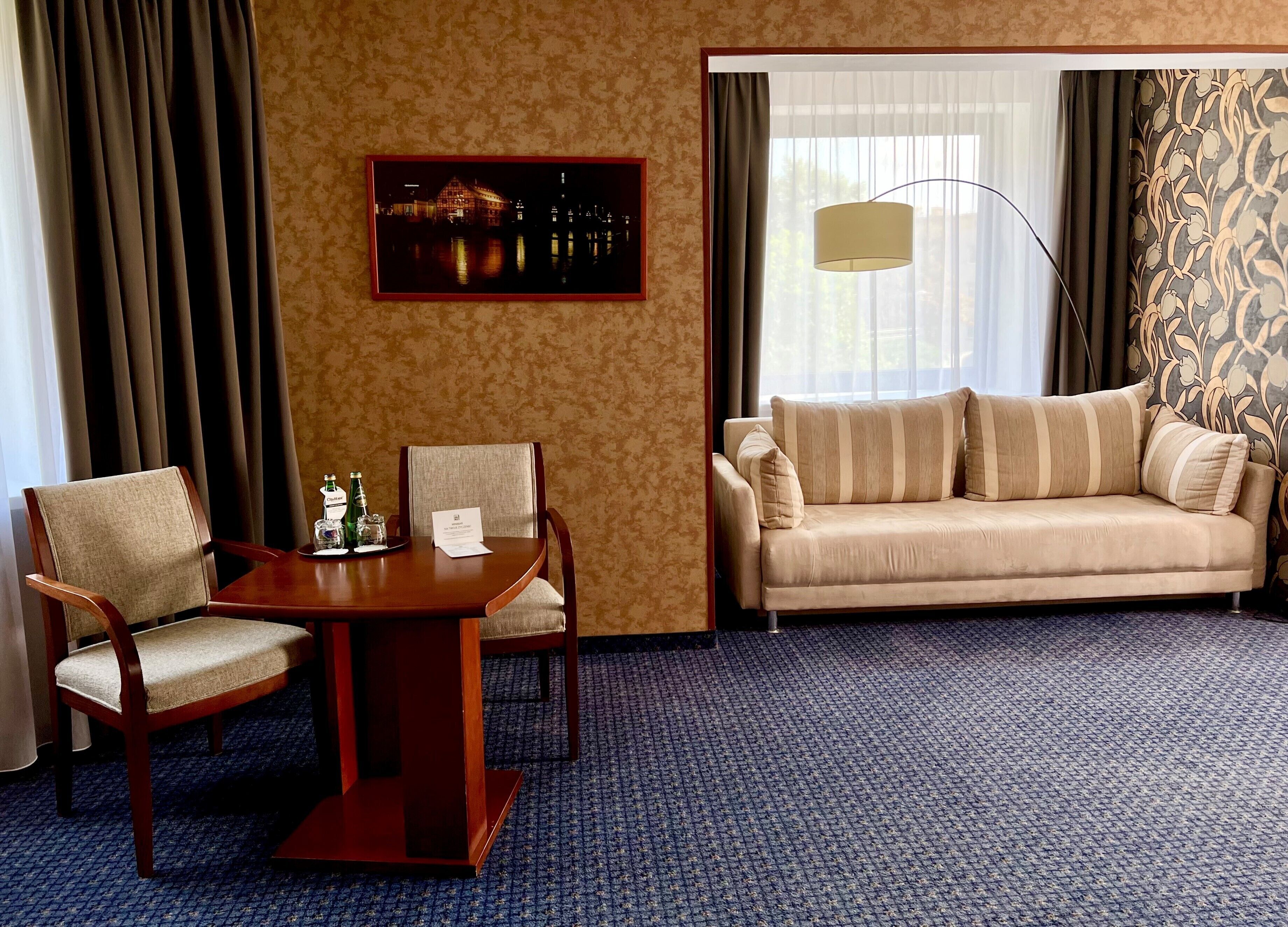 Habitación de hotel con una mesa de comedor de madera, dos sillas, un sofá, un cuadro en la pared y un ventanal con cortinas.