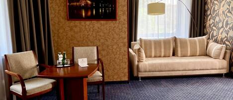 Habitación de hotel con una mesa de comedor de madera, dos sillas, un sofá, un cuadro en la pared y un ventanal con cortinas.