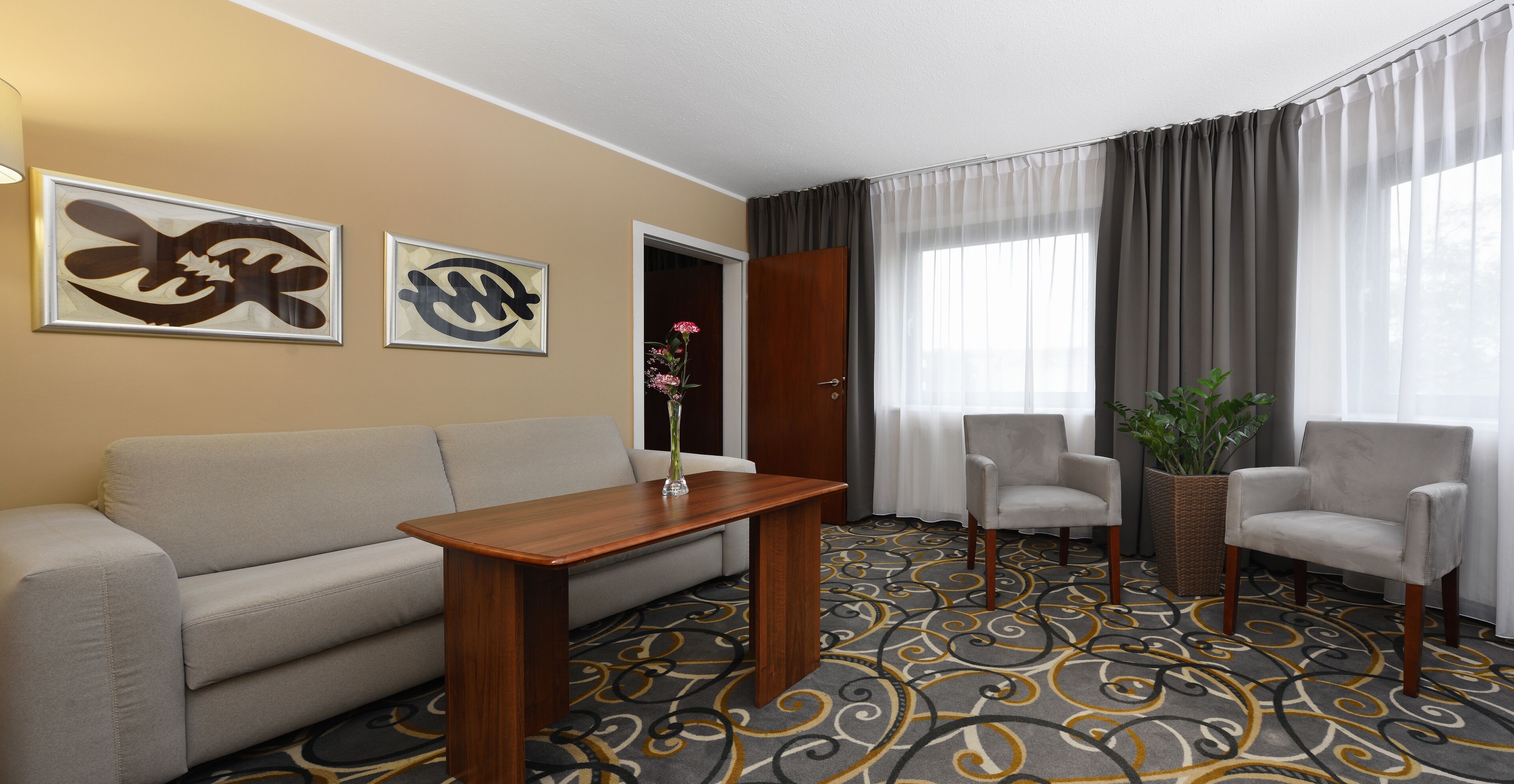 Quarto de hotel com sofá, mesa de madeira, duas poltronas, tapete estampado e duas obras de arte abstratas nas paredes.