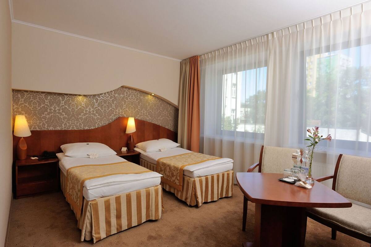 Habitación de hotel con dos camas, cabecero de madera, una mesa con sillas y una ventana con cortinas.
