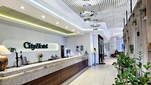 Zona de recepción de hotel con mostrador, lámpara, cartel en la pared y plantas en macetas.