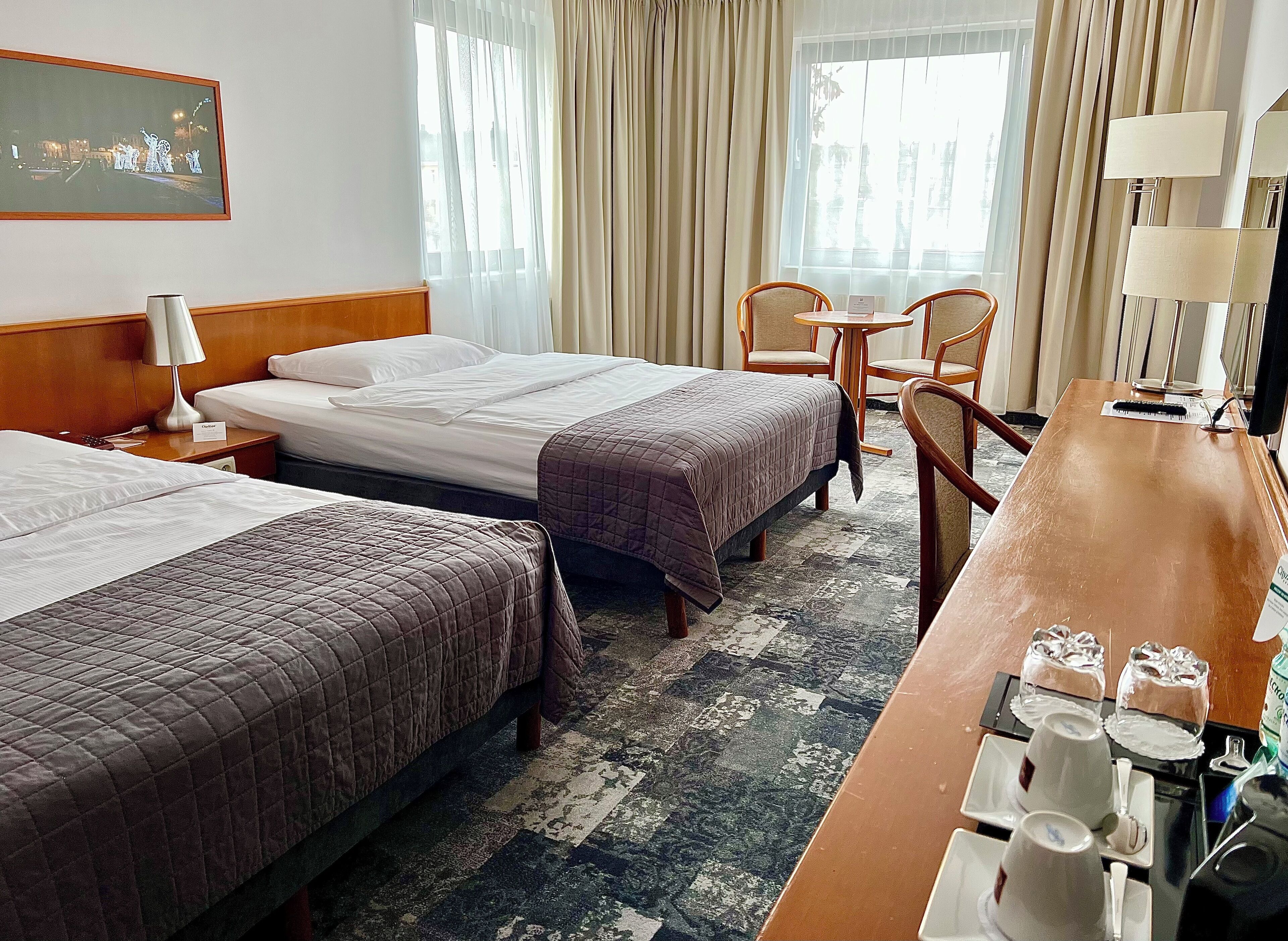 A hotel room with two beds, a desk with a chair, a small table, and a large window with curtains.