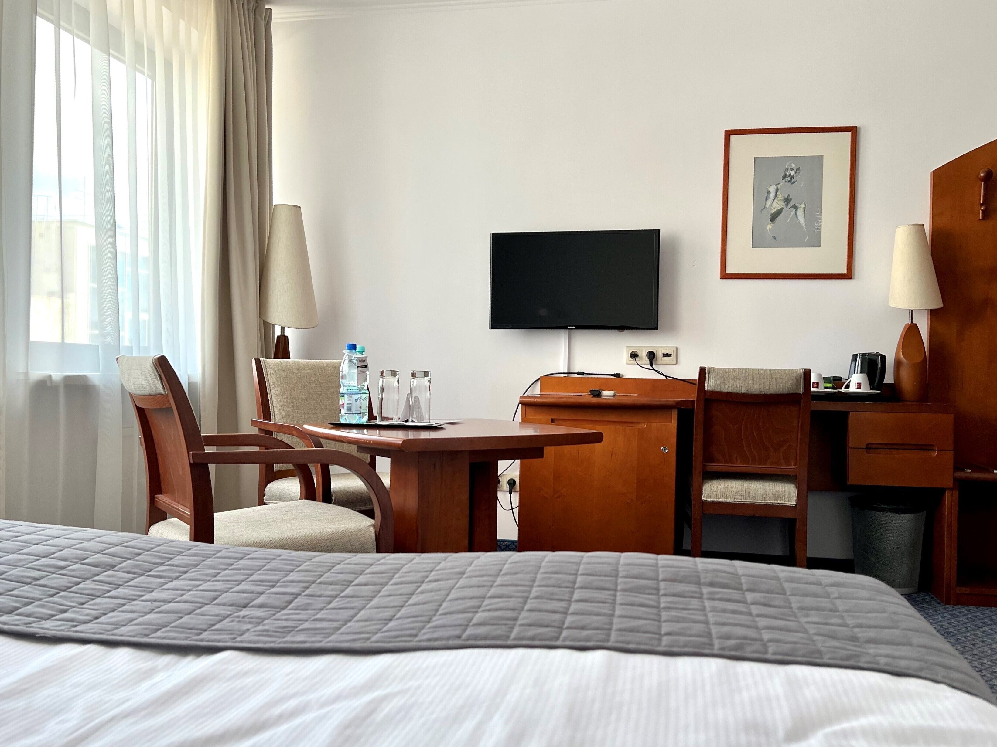 A hotel room with a bed, a desk with a television, chairs, a lamp, a framed picture, and a water bottle.