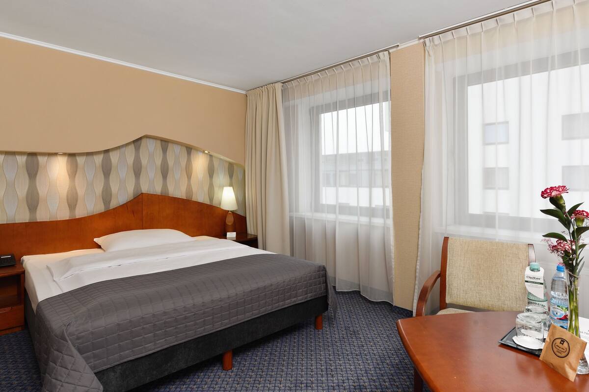 Habitación de hotel con cama, mesita de noche, lámpara, silla, mesa con un florero y ventana con cortinas.