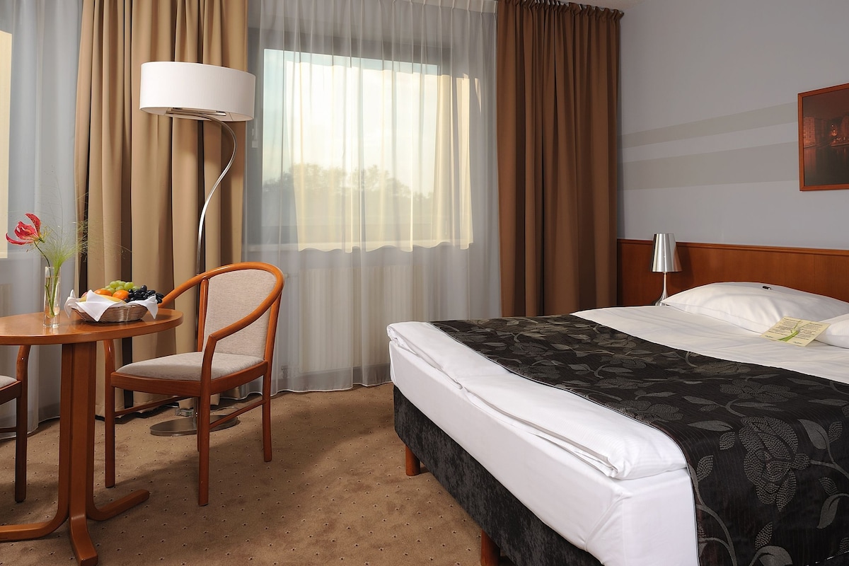 A hotel room with a bed, two chairs, a table with a basket of fruit, a lamp, and a painting on the wall.