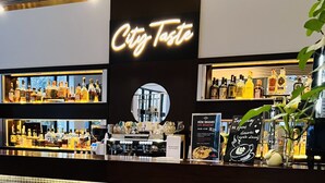 Zona de bar con un letrero de neón que dice "City Taste" y estantes llenos de botellas de licor.