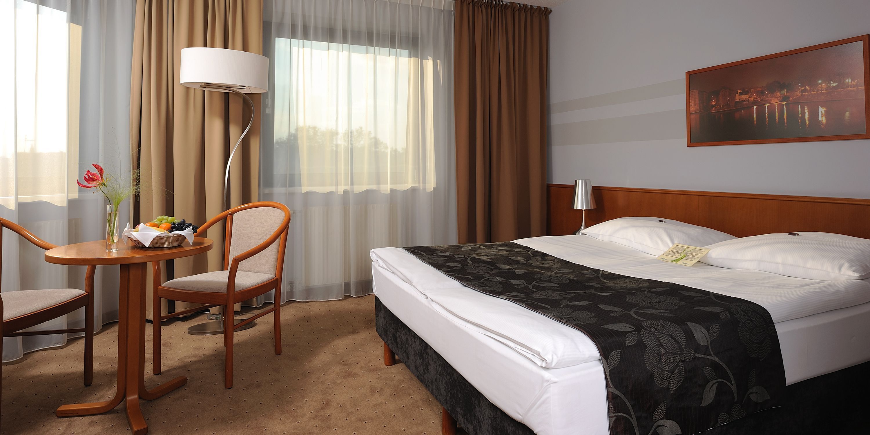 A hotel room with a bed, a desk with a chair, a lamp, and a window with curtains.