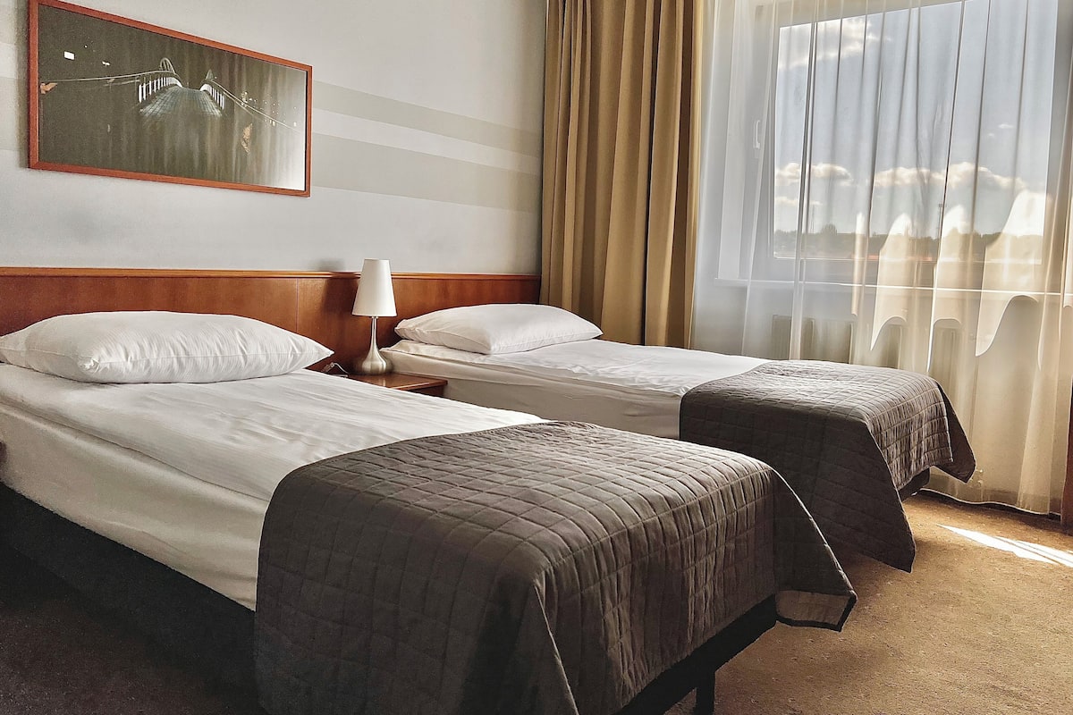 Habitación de hotel con dos camas, una mesita de noche, una lámpara, un cuadro en la pared y una ventana con cortinas.