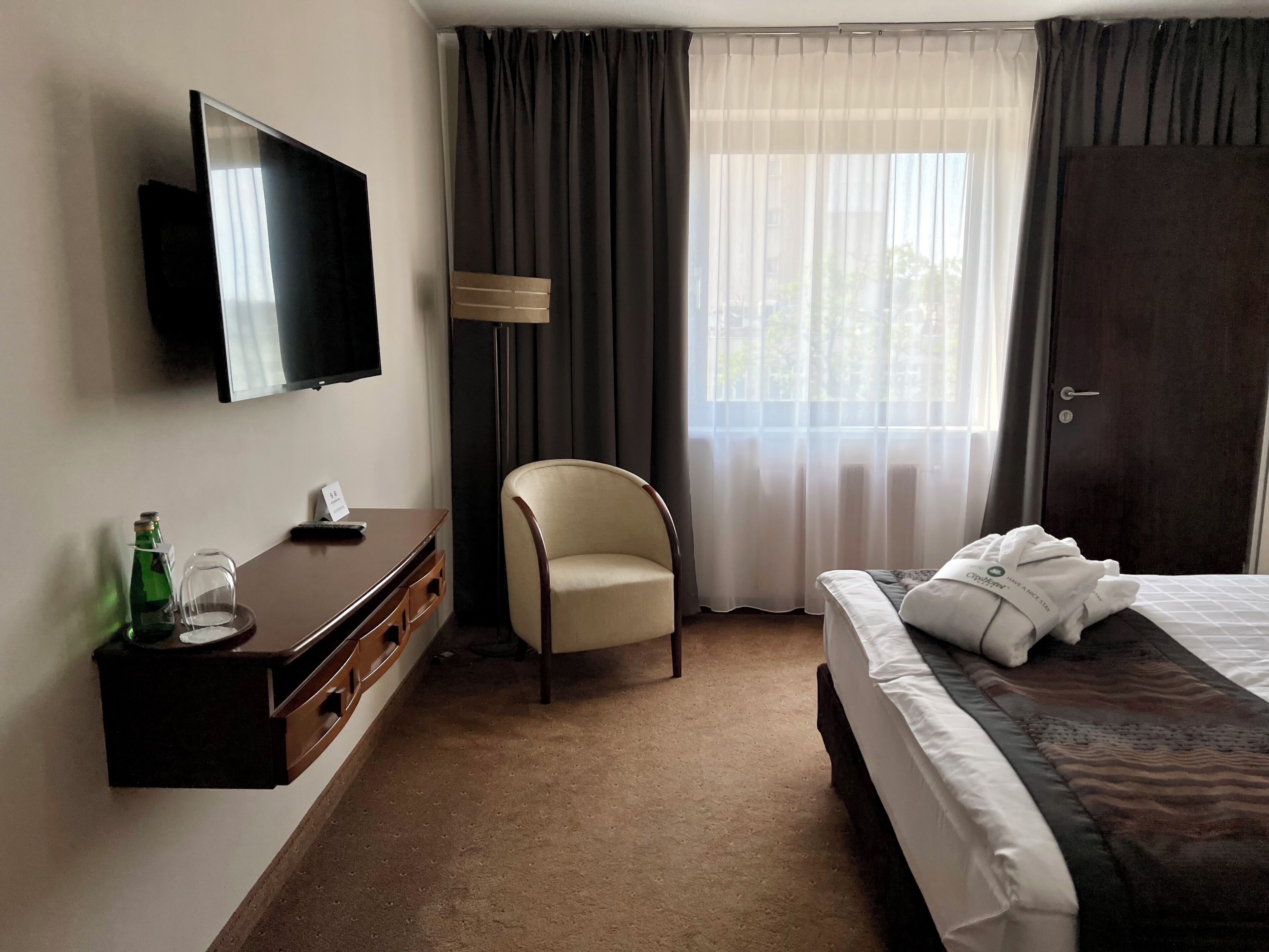 A hotel room with a bed, a chair, a TV mounted on the wall, a dresser, and a window with curtains.