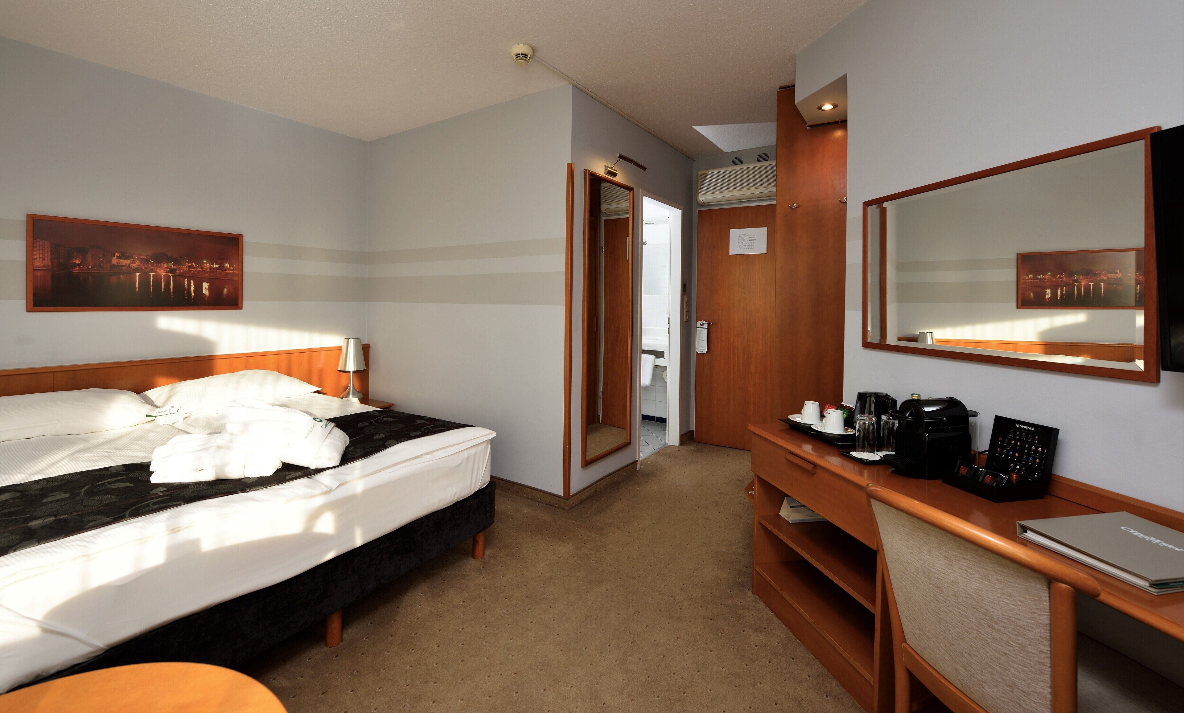 A hotel room with a bed, a desk with a coffee maker, a mirror, and a picture of a cityscape.