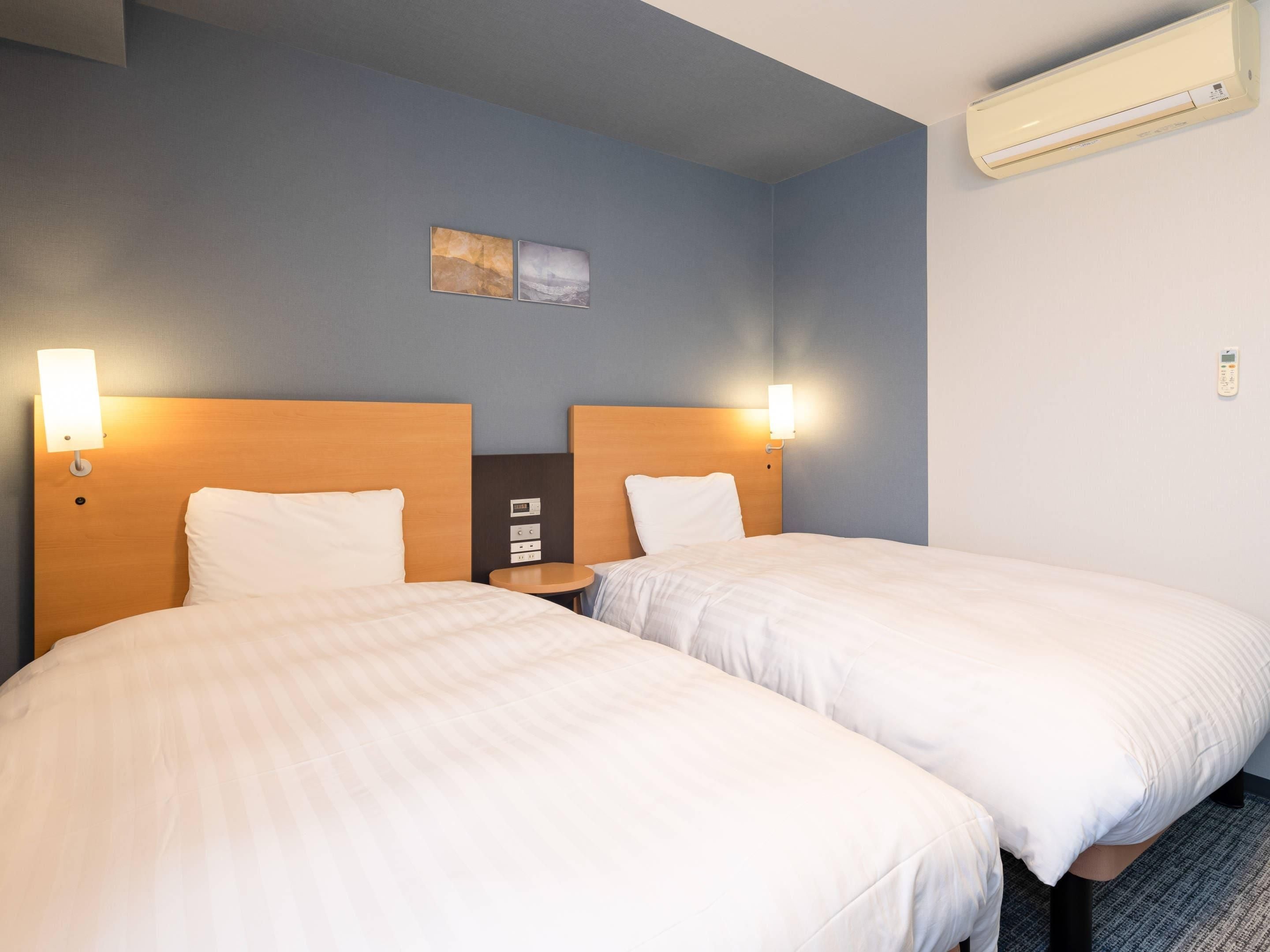 A hotel room with two beds, a nightstand, a wall-mounted air conditioner, and a small artwork above the beds.