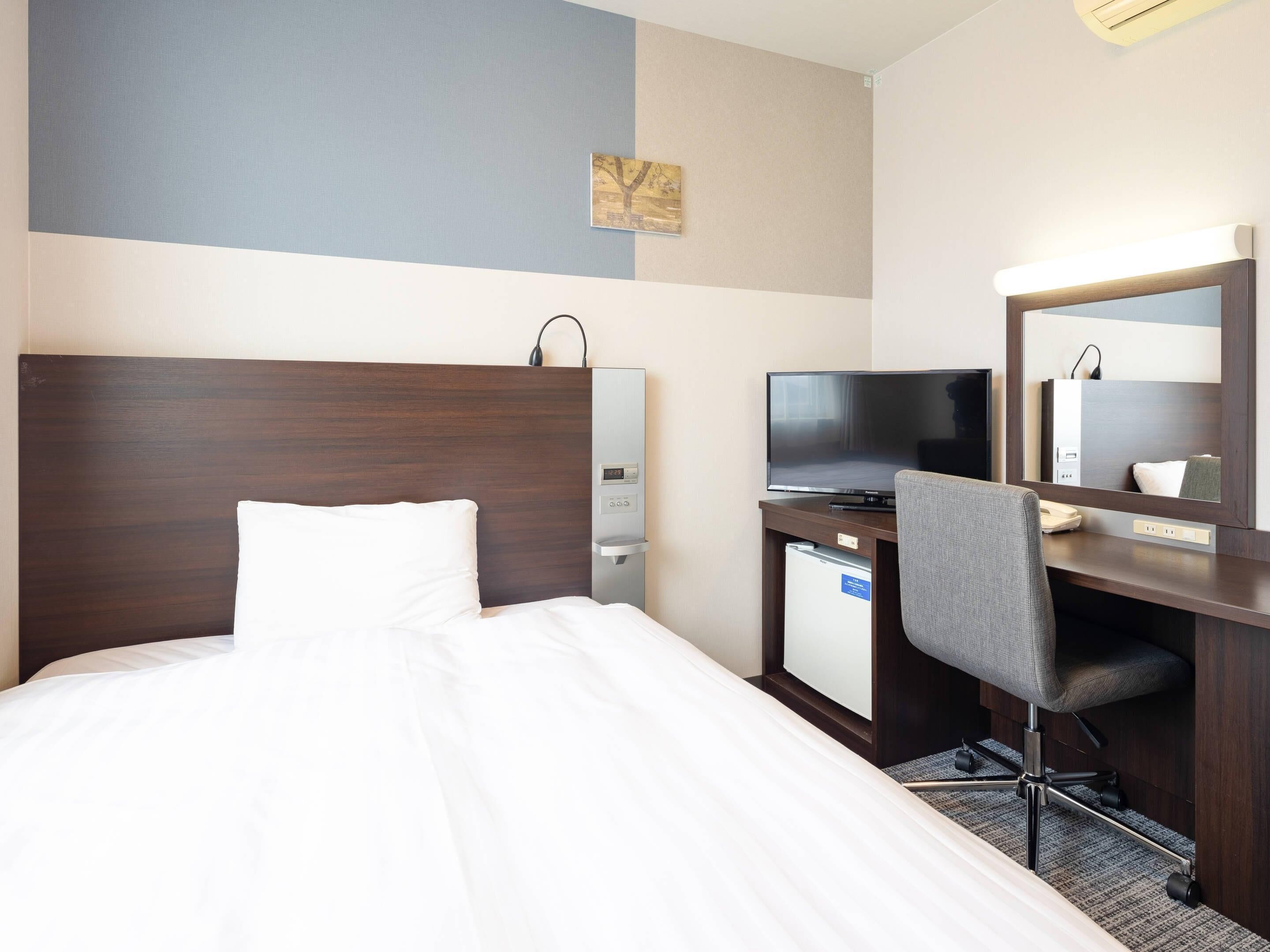 A hotel room with a bed, a desk with a computer, a chair, a TV, and a small refrigerator.