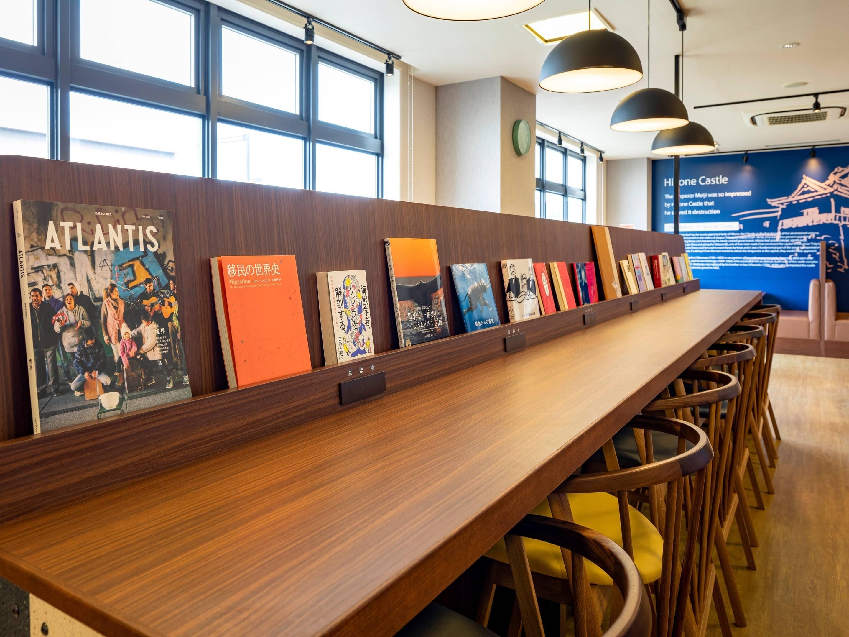 A long wooden table with books and chairs in a well-lit room with large windows and a blue informational display in the background.