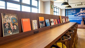 A long wooden table with books and chairs in a well-lit room with large windows and a blue informational display in the background.