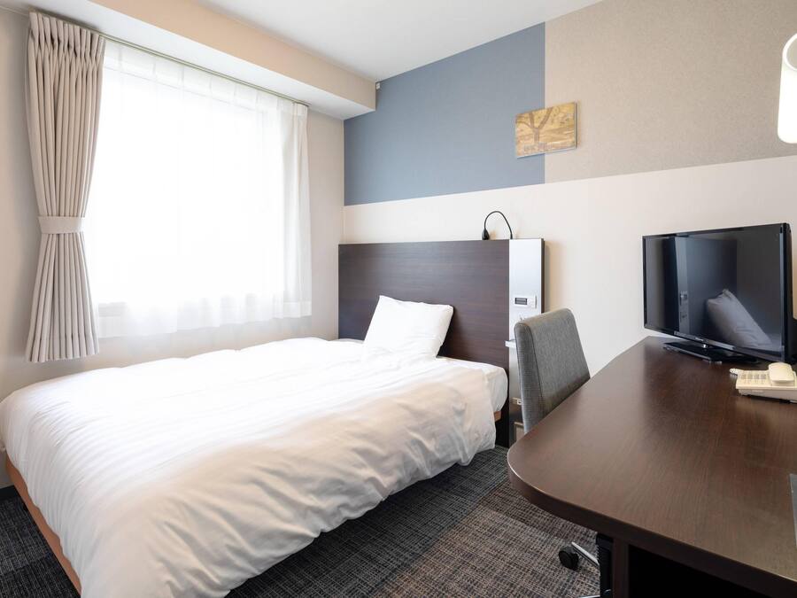 A hotel room with a bed, a desk with a television, a chair, and a window with curtains.