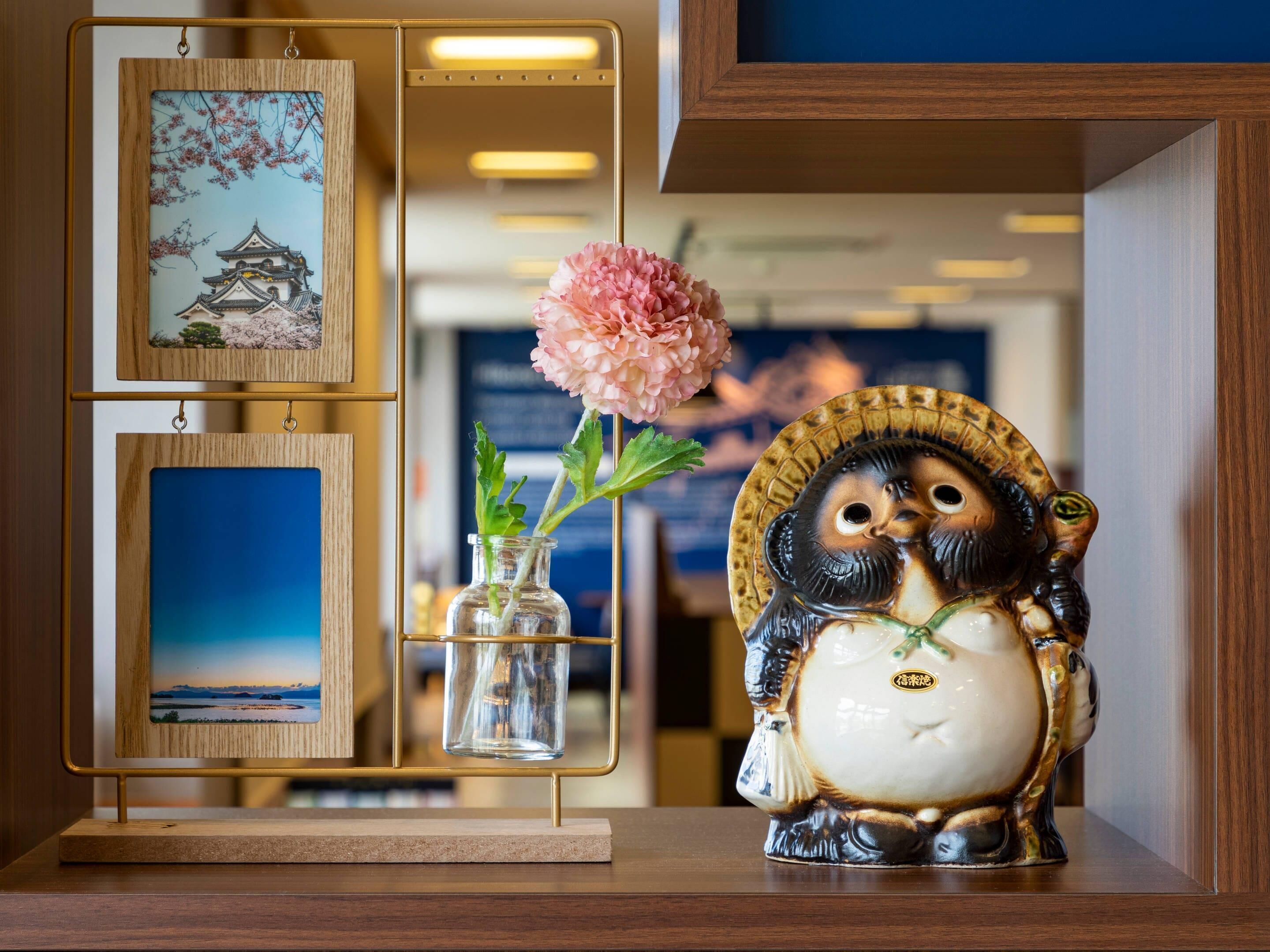 Una repisa decorativa con una pintura tradicional de un edificio japonés, un florero con una flor rosa y una figurita de cerámica de un búho.