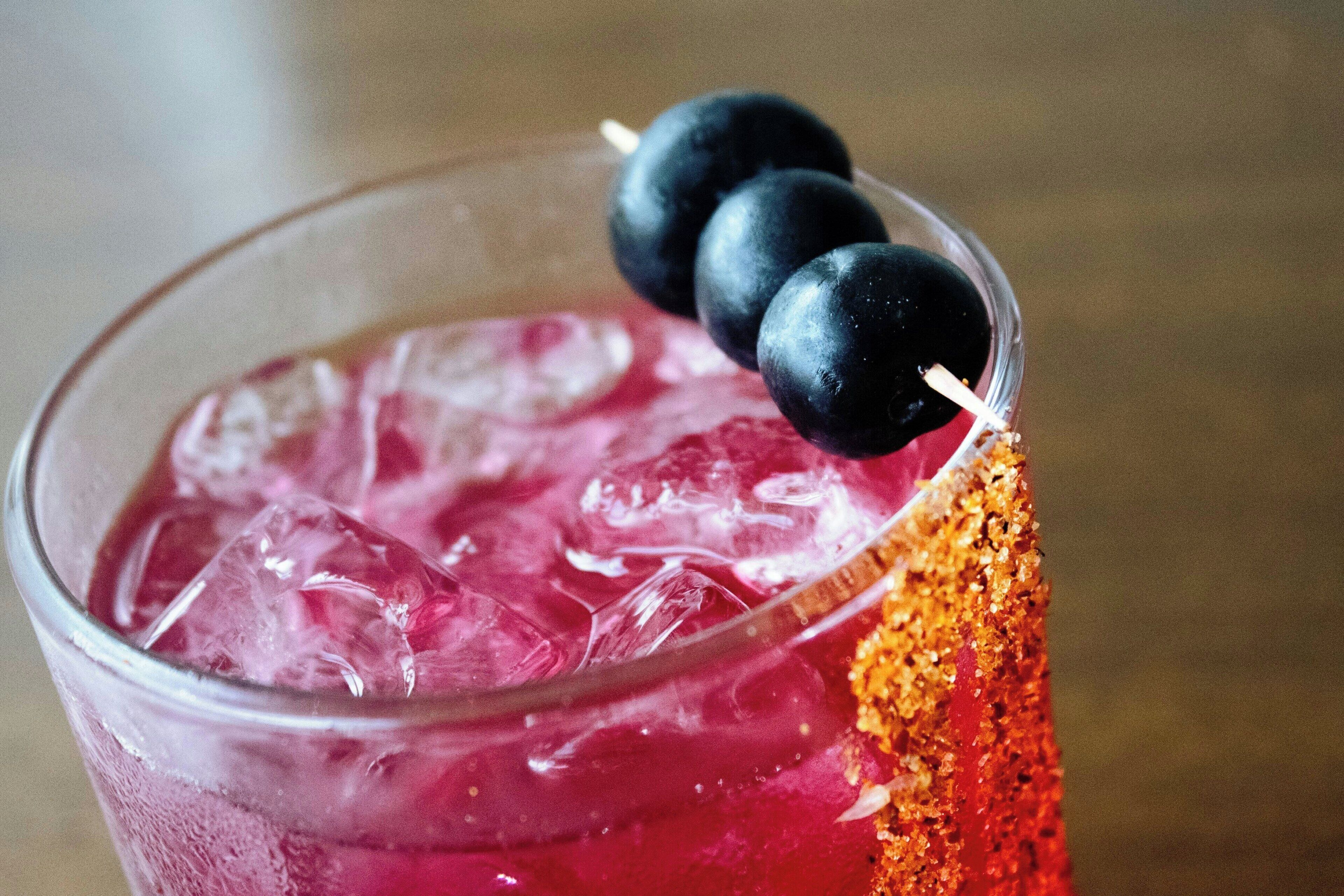 Een glas roze drank met ijsblokjes, garnering van bosbessen en een beetje oranje poeder.