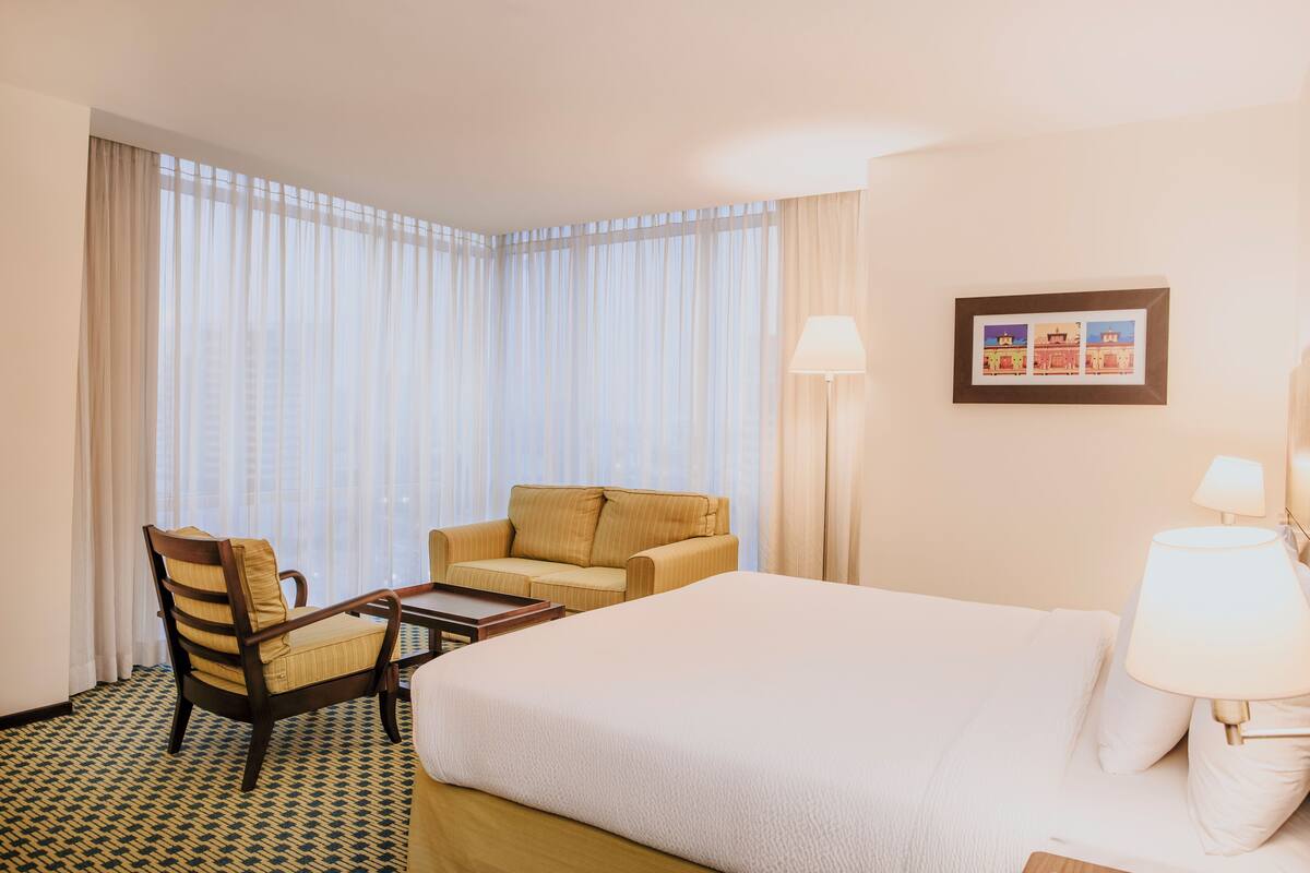 Een hotelkamer met een groot bed, een houten stoel, een kleine tafel en een bank. Er is een raam met dunne gordijnen en een lijst met een afbeelding aan de muur.