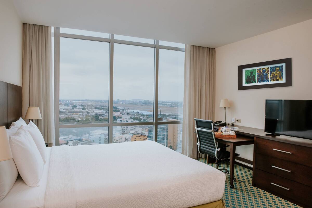 Une chambre d’hôtel avec un grand lit, un bureau avec une chaise, une télévision et une vue sur la ville grâce à de grandes fenêtres.