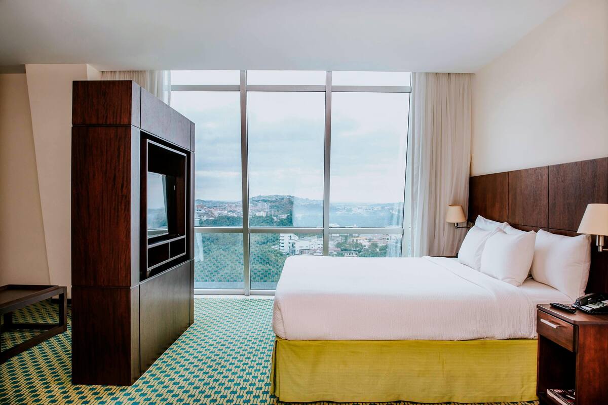 Une chambre d’hôtel avec un grand lit, une télévision, une table de chevet et une vue sur la ville grâce à de grandes fenêtres.