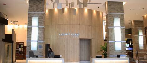 Een moderne hotel lobby met een receptiebalie, een 'Courtyard by Marriott' bord en een grote plant.